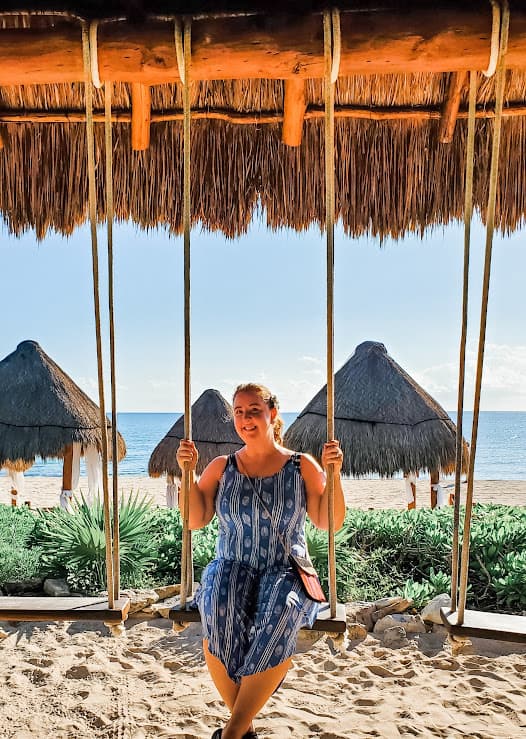 Laura in a blue dress sitting on a wooden swing outdoors on the beach with the ocean visible behind her