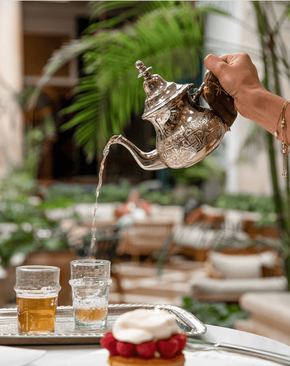 View of a hand pouring mint tea into a glass from a shiny teapot indoors with plants and other tables visible in the background