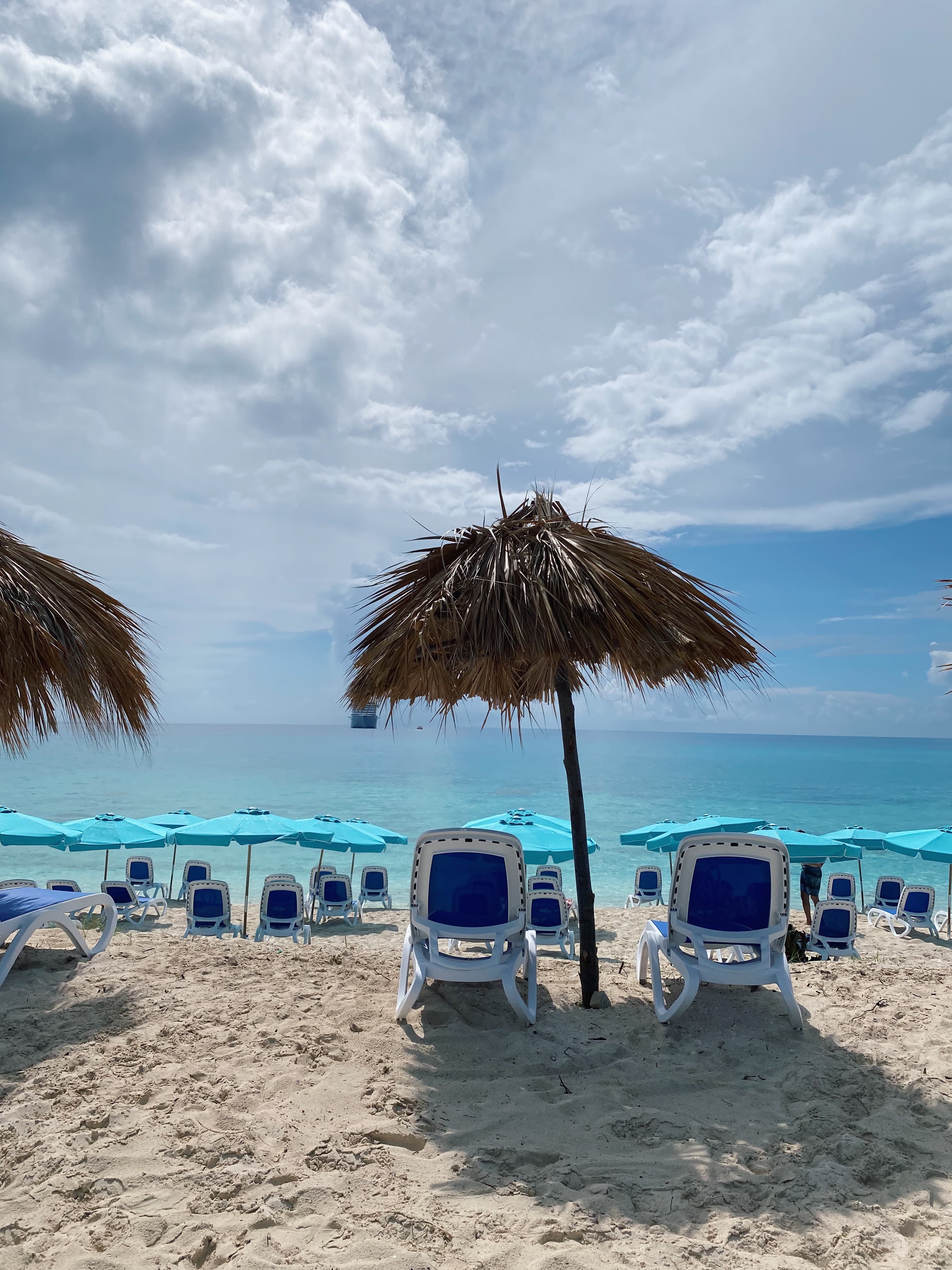 Gentle waves lap the shore of a beach lined with loungers and grass umbrellas that sway in the breeze on a sunny day.