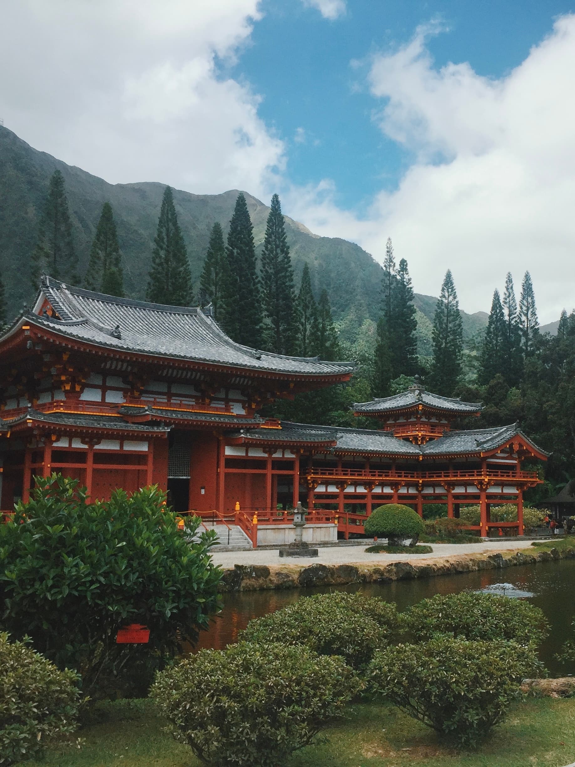 Historical building with a pitched roof stands against a backdrop of evergreen trees and hills on a cloudy day.