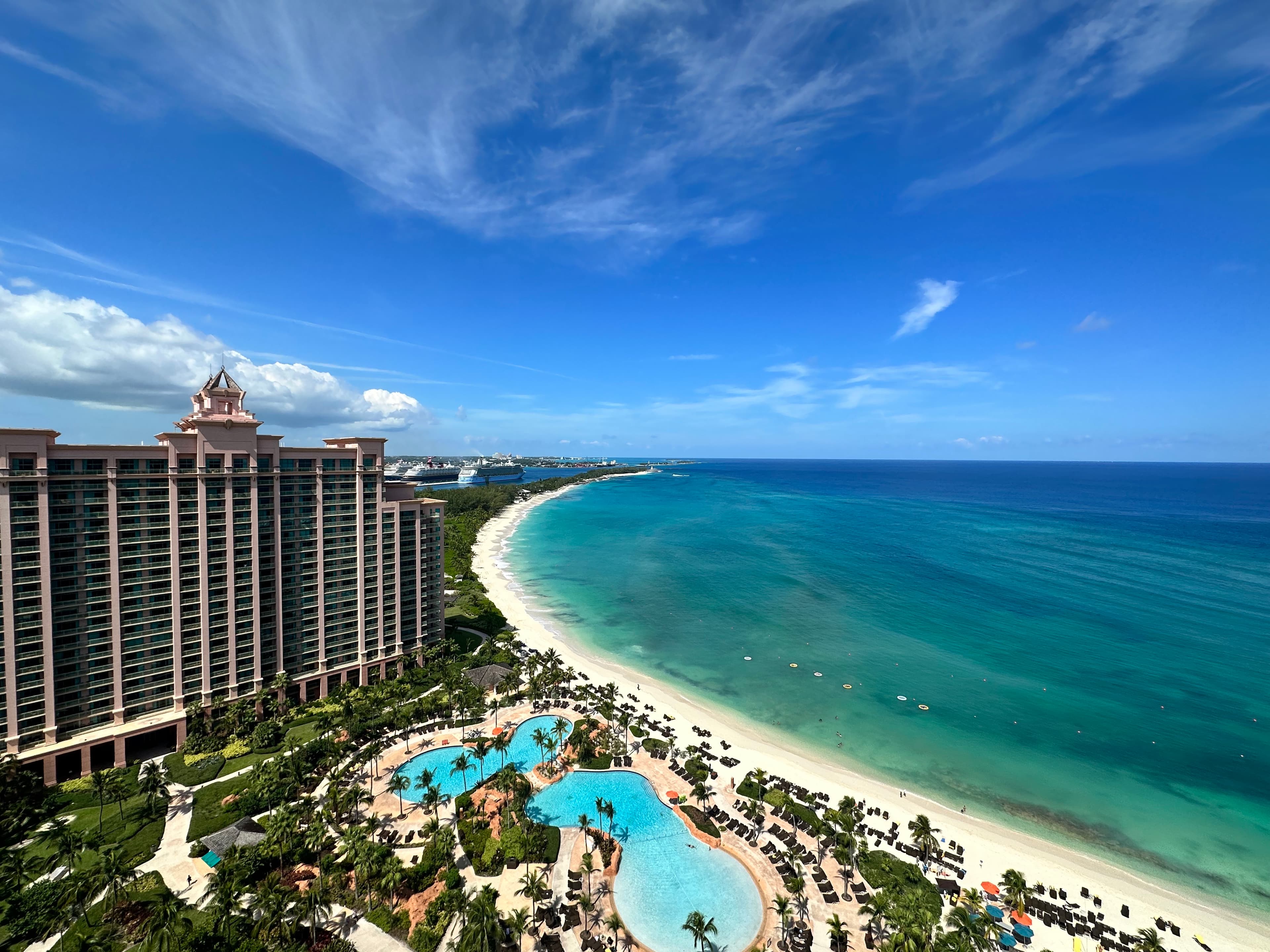 Aerial view of a beachfront resort with a large pool area, tall hotel building, and a long stretch of white sandy beach meeting the turquoise ocean under a blue sky with scattered clouds.