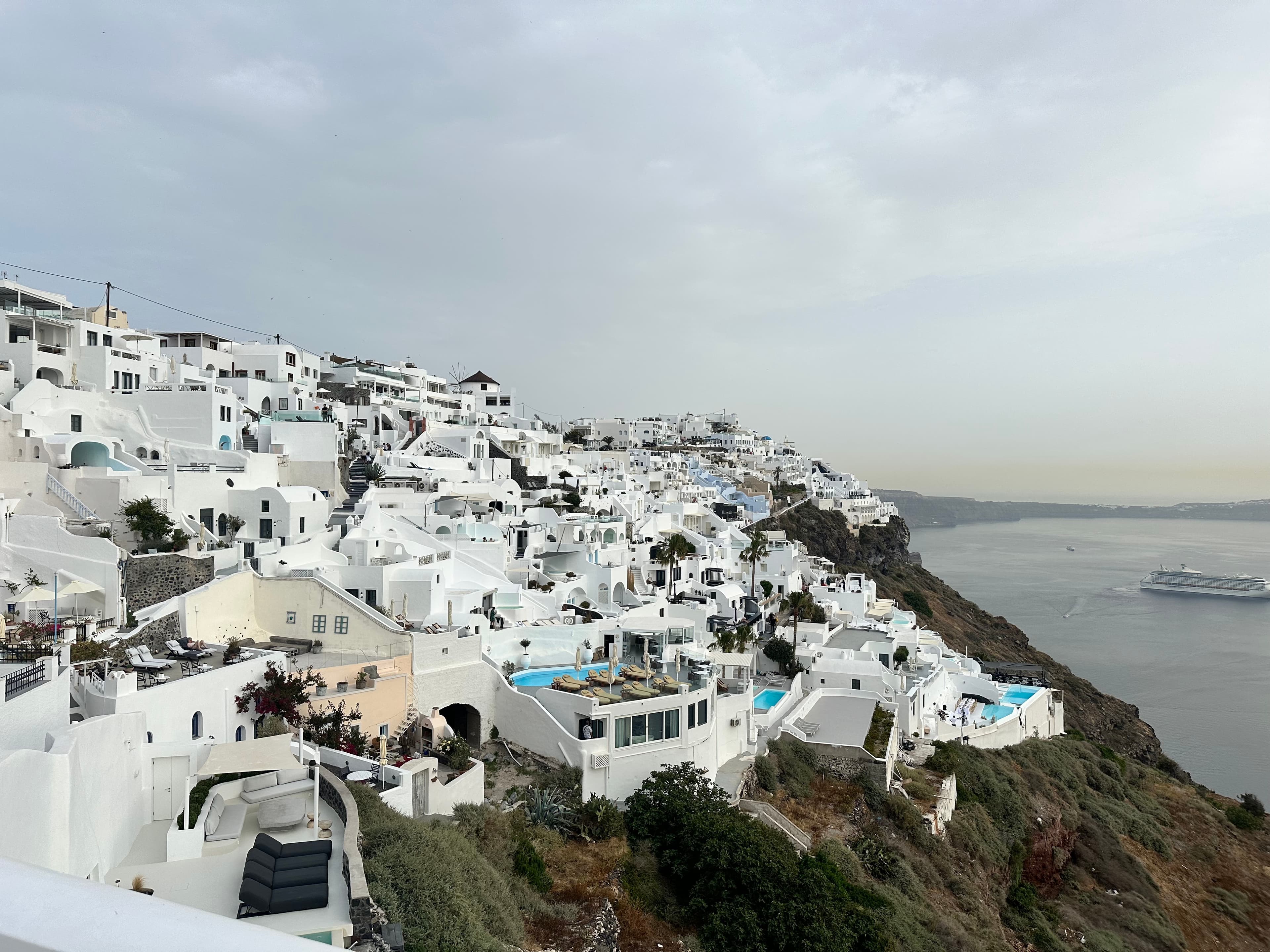 Panoramic view of Oia village from hilltop, overlooking white buildings and blue domes against the Aegean Sea.