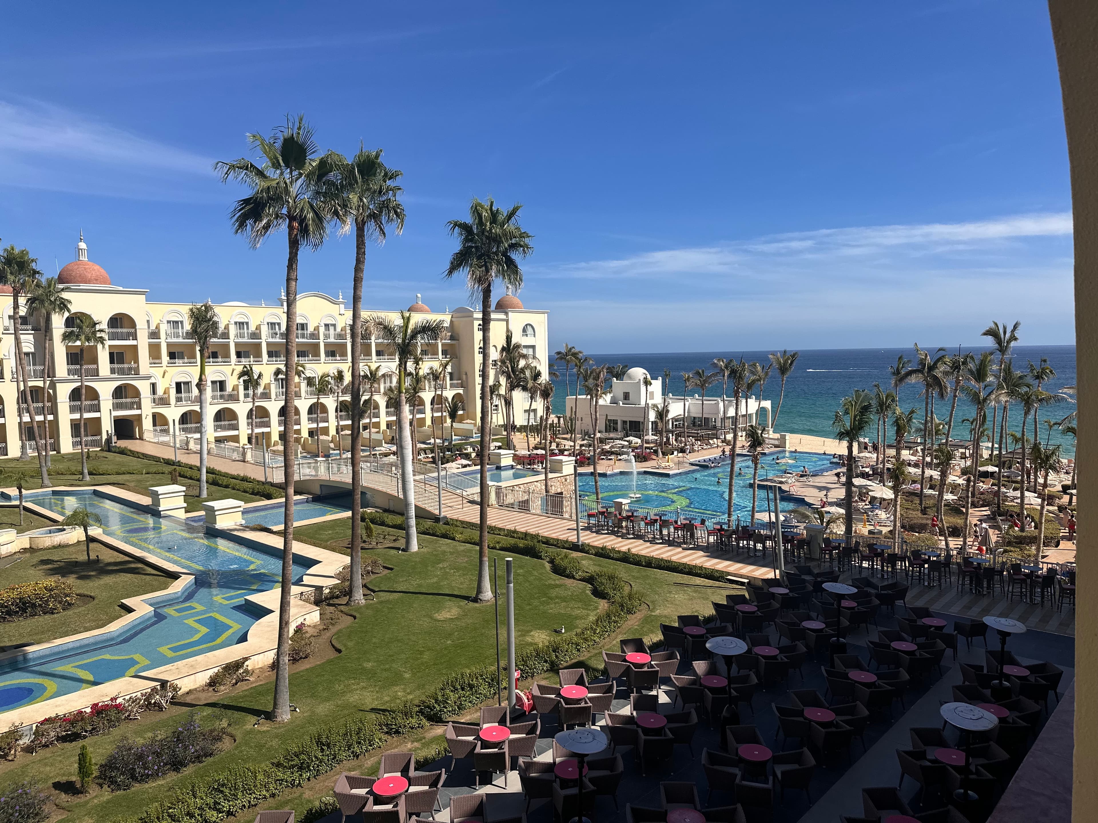 View of a beachfront resort featuring a large building with arched windows, multiple pools, palm trees, and a patio area with tables and chairs, overlooking the ocean under a clear blue sky.