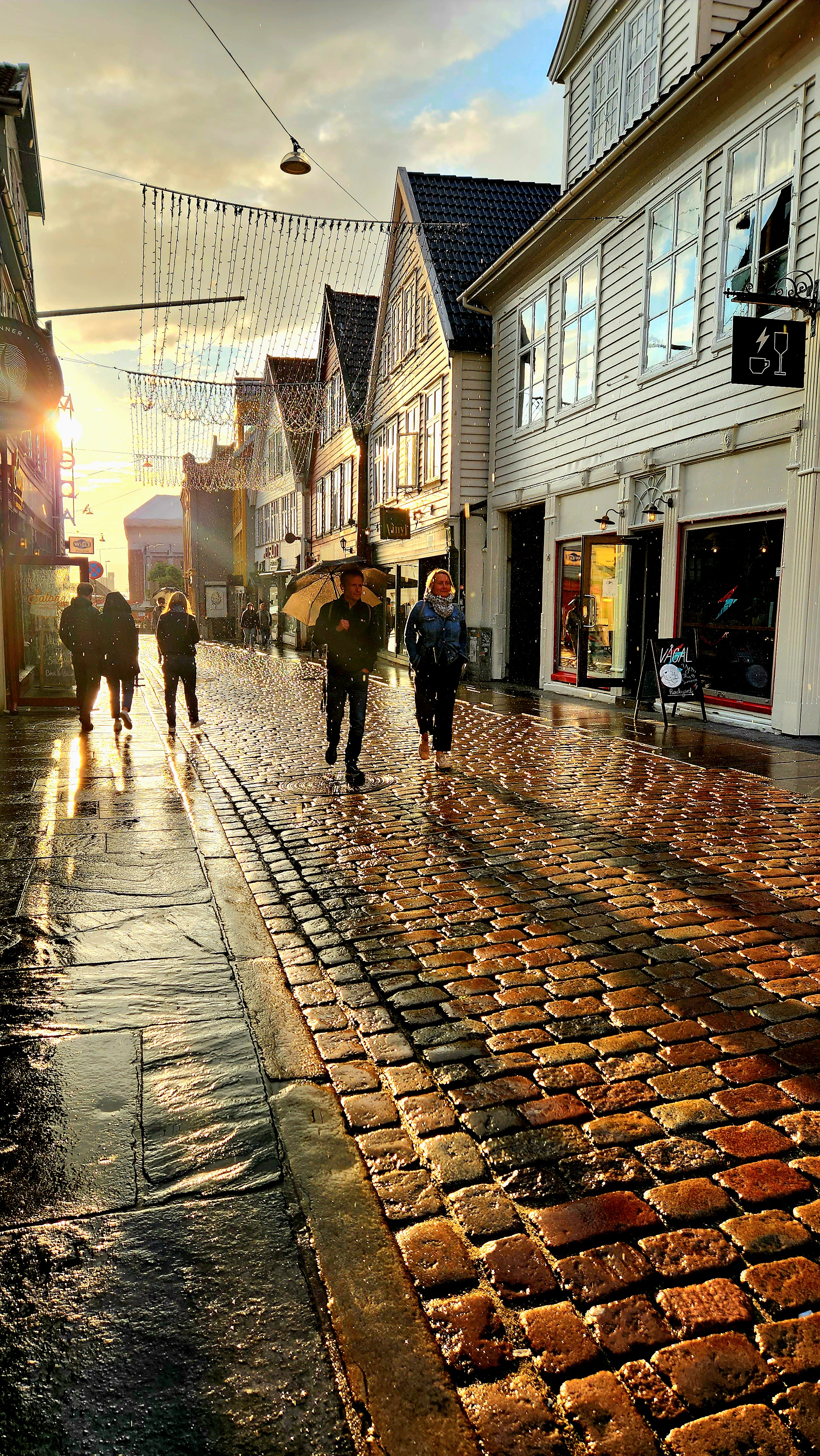 A view of cobblestone streets and people walking around. The sun in shining through the clouds in the background.