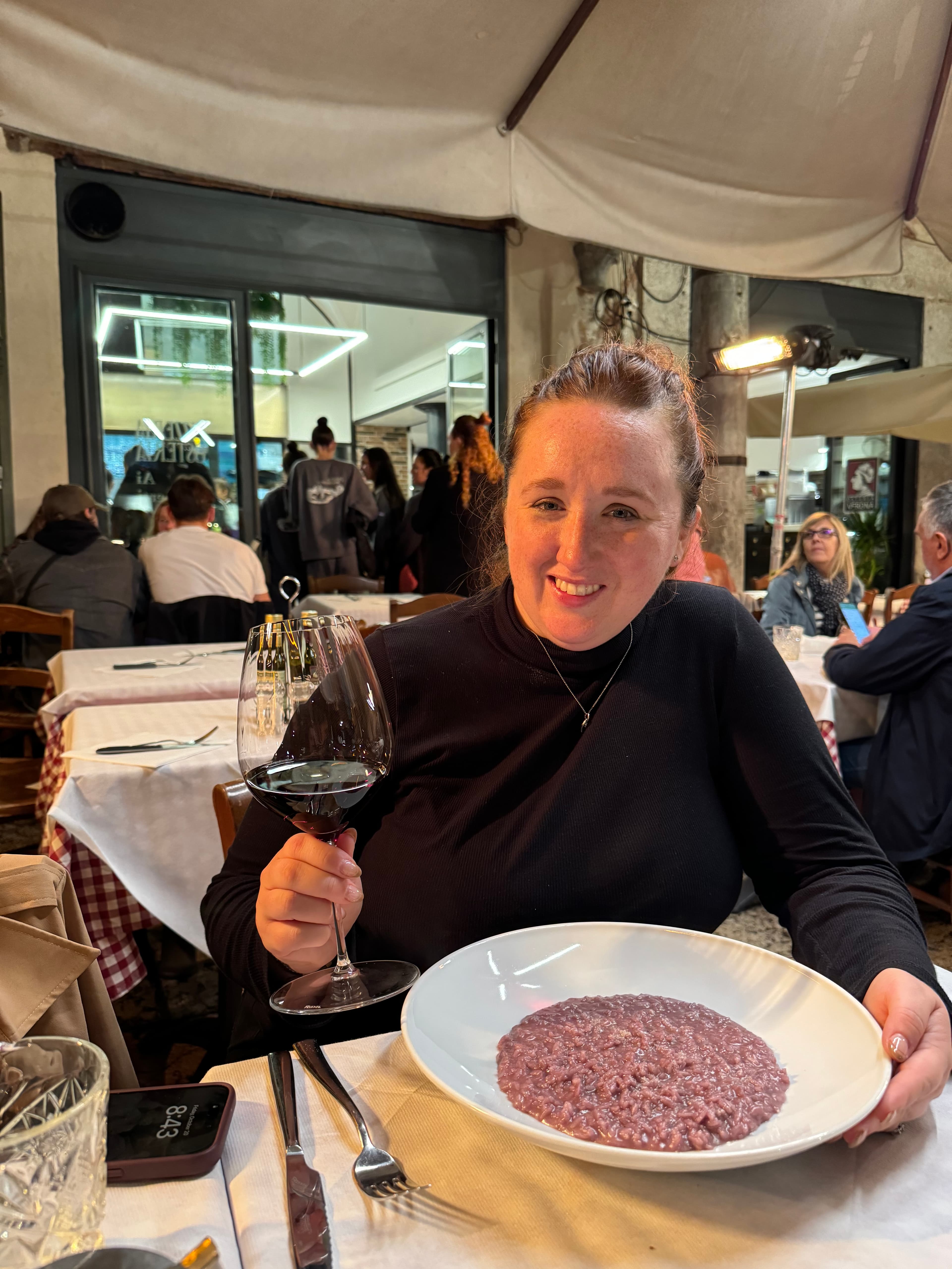 Briana holding a glass of red wine at a restaurant table in Italy with a plate of risotto in front of her