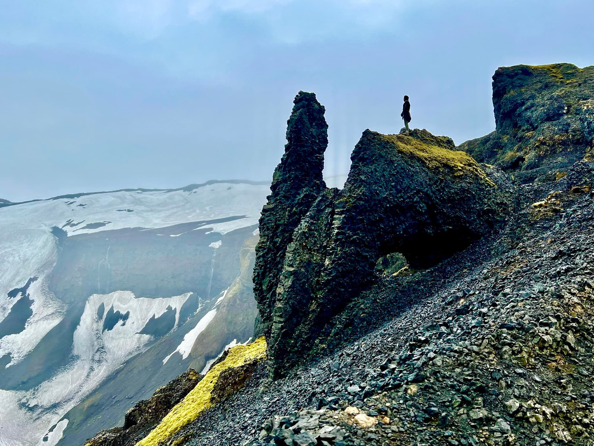 View of Jonathan standing on a dramatic black rock cliffside overlooking glaciers under cloudy skies
