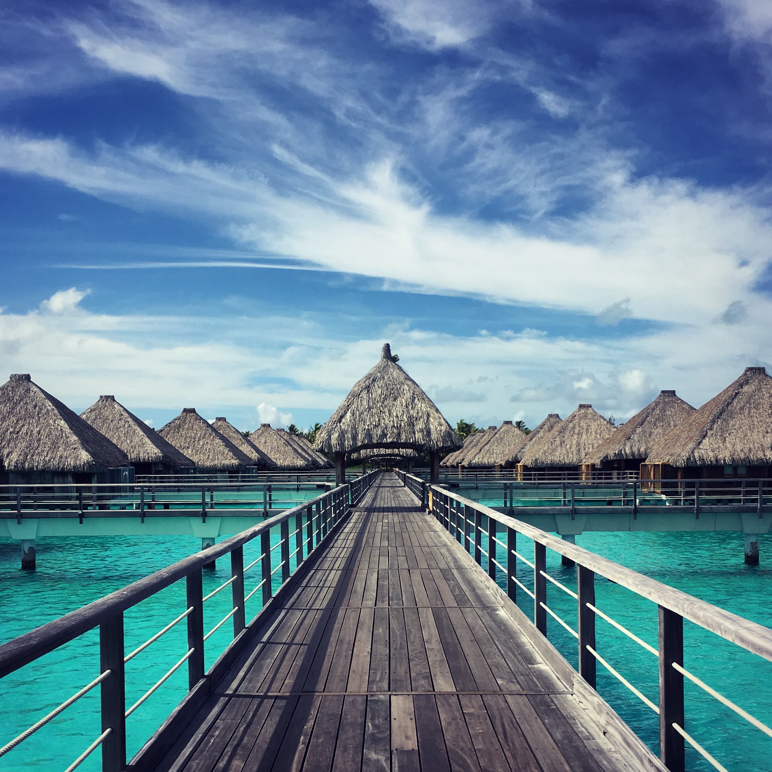 View of a narrow wooden pathway over turquoise waters leading to several bungalows