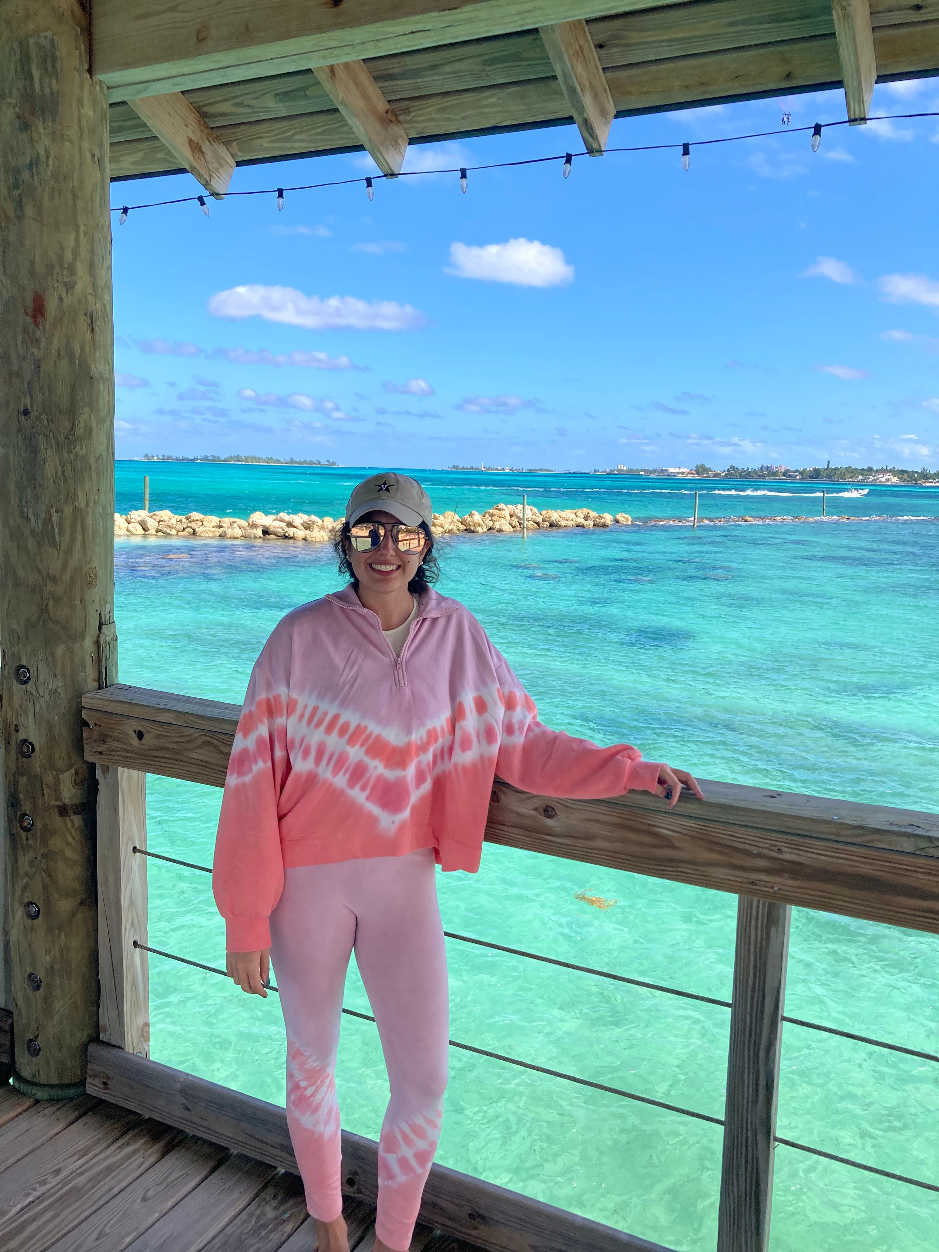 Michelle in sunglasses and a baseball cap holding on to a railing overlooking a turquoise sea and clear skies