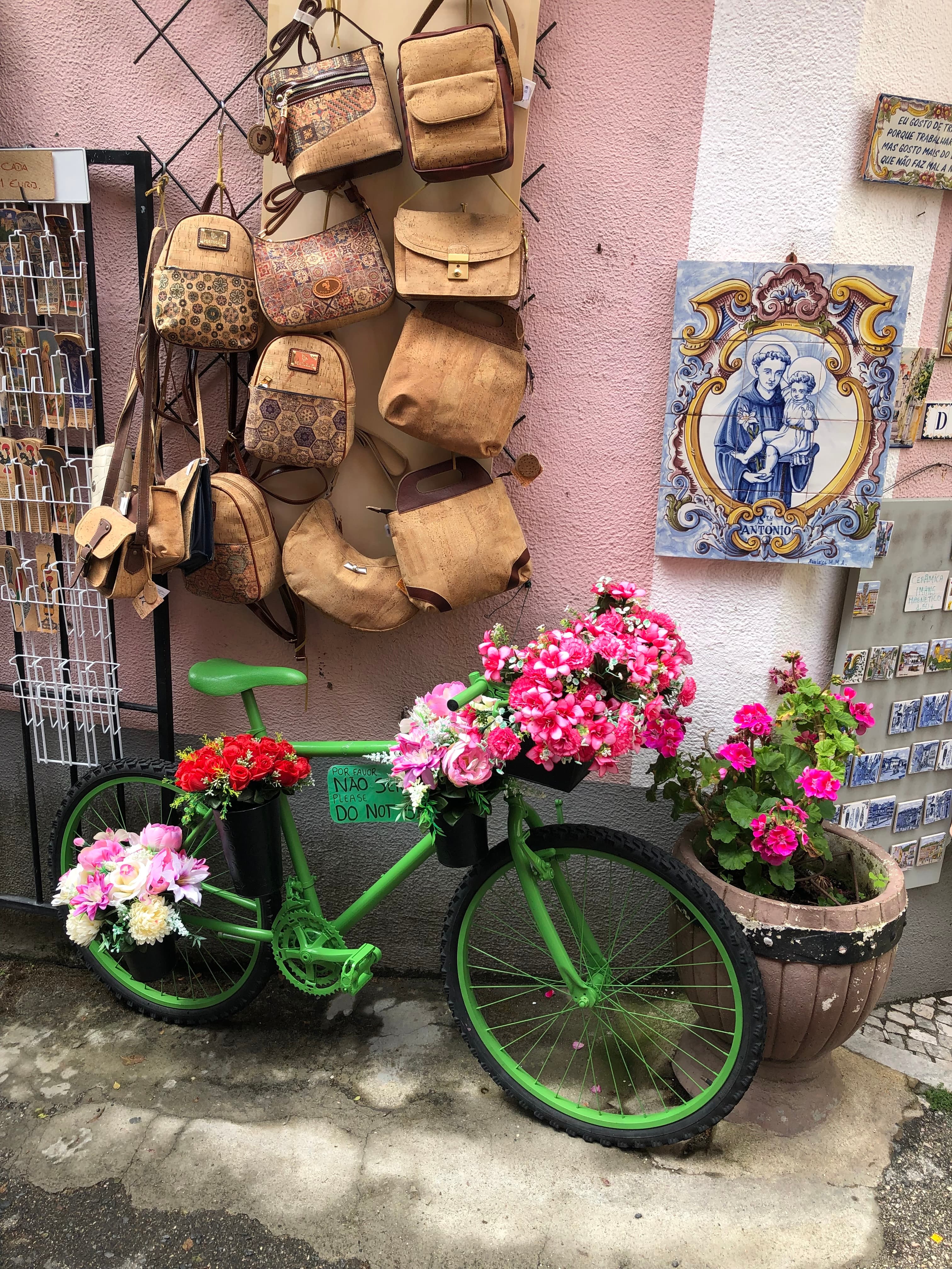A green bike with pink flowers in a basket parked in front of a wall with bags on display