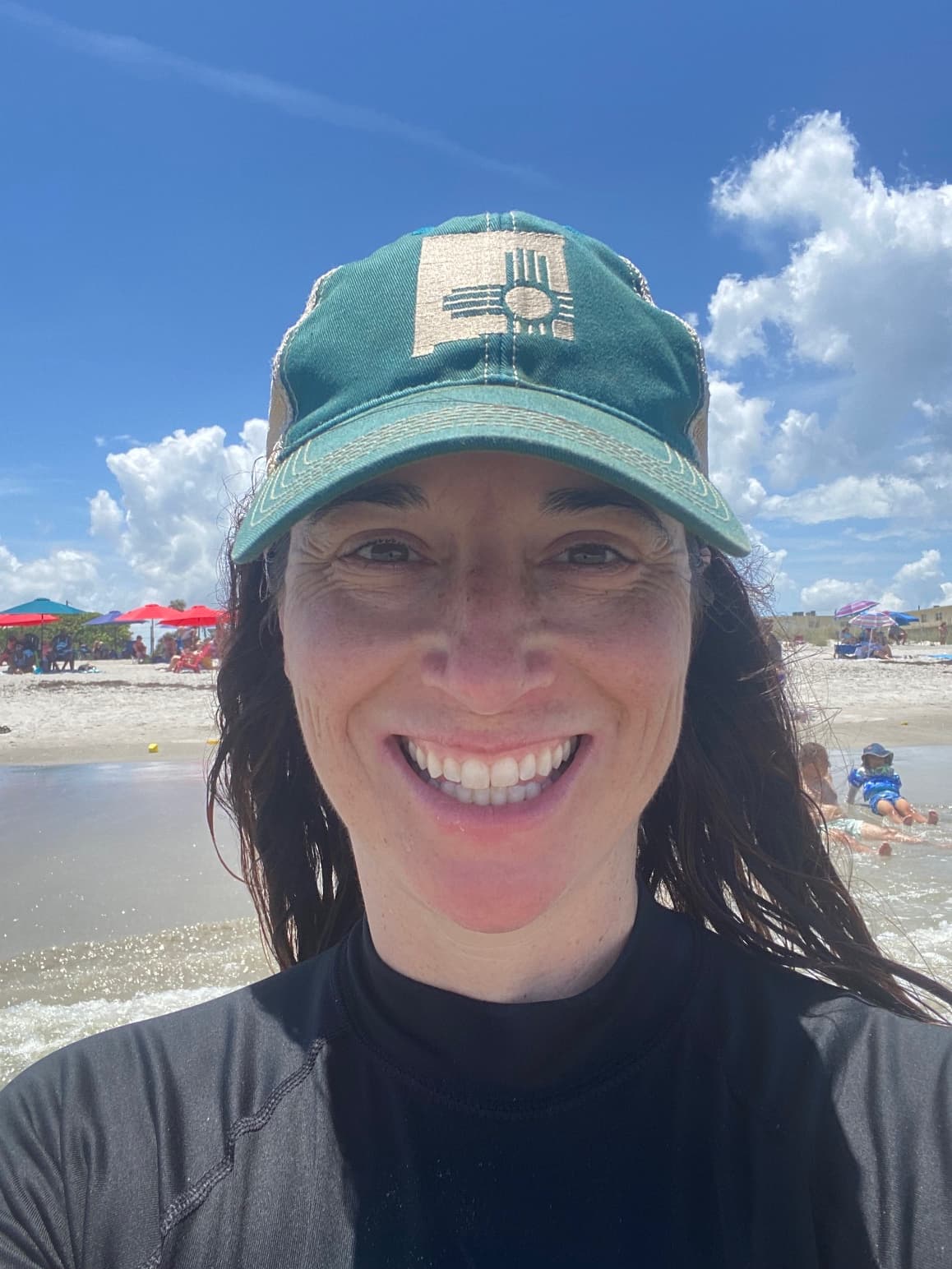 Renee in a green baseball cap and wetsuit taking a selfie on the beach on sunny day