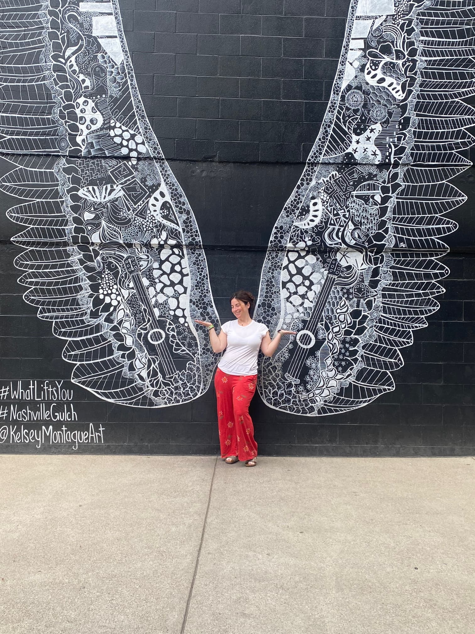 Advisor in red pants standing in front of a black brick wall with a painting of large white wings behind her