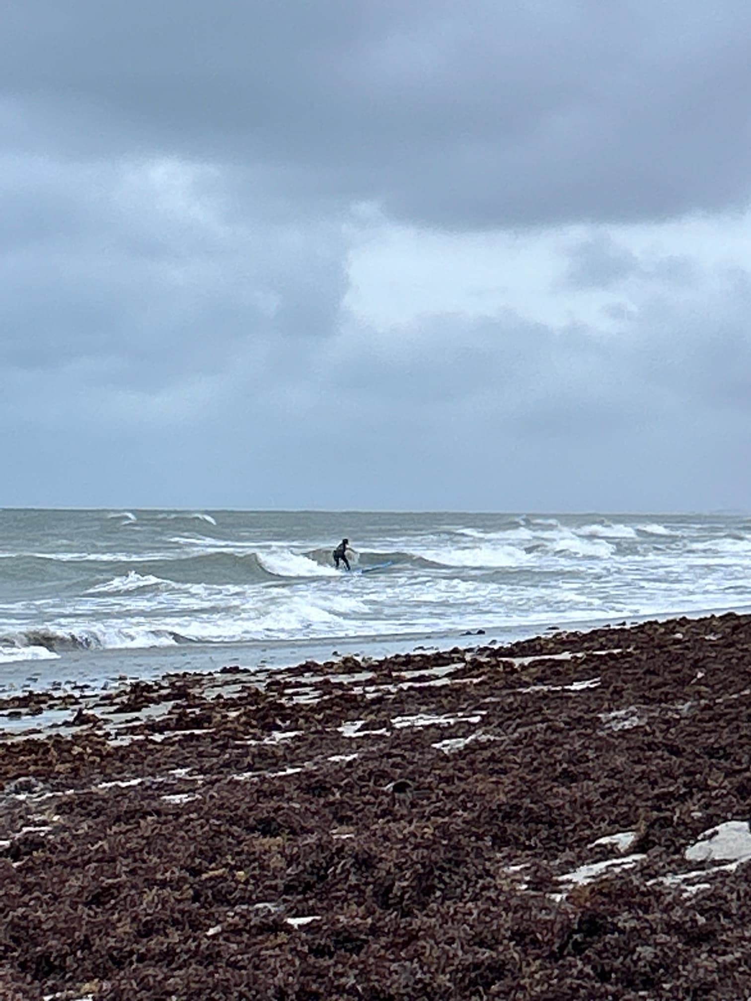 View of a winter coastline with a surfer on a small wave under cloudy skies