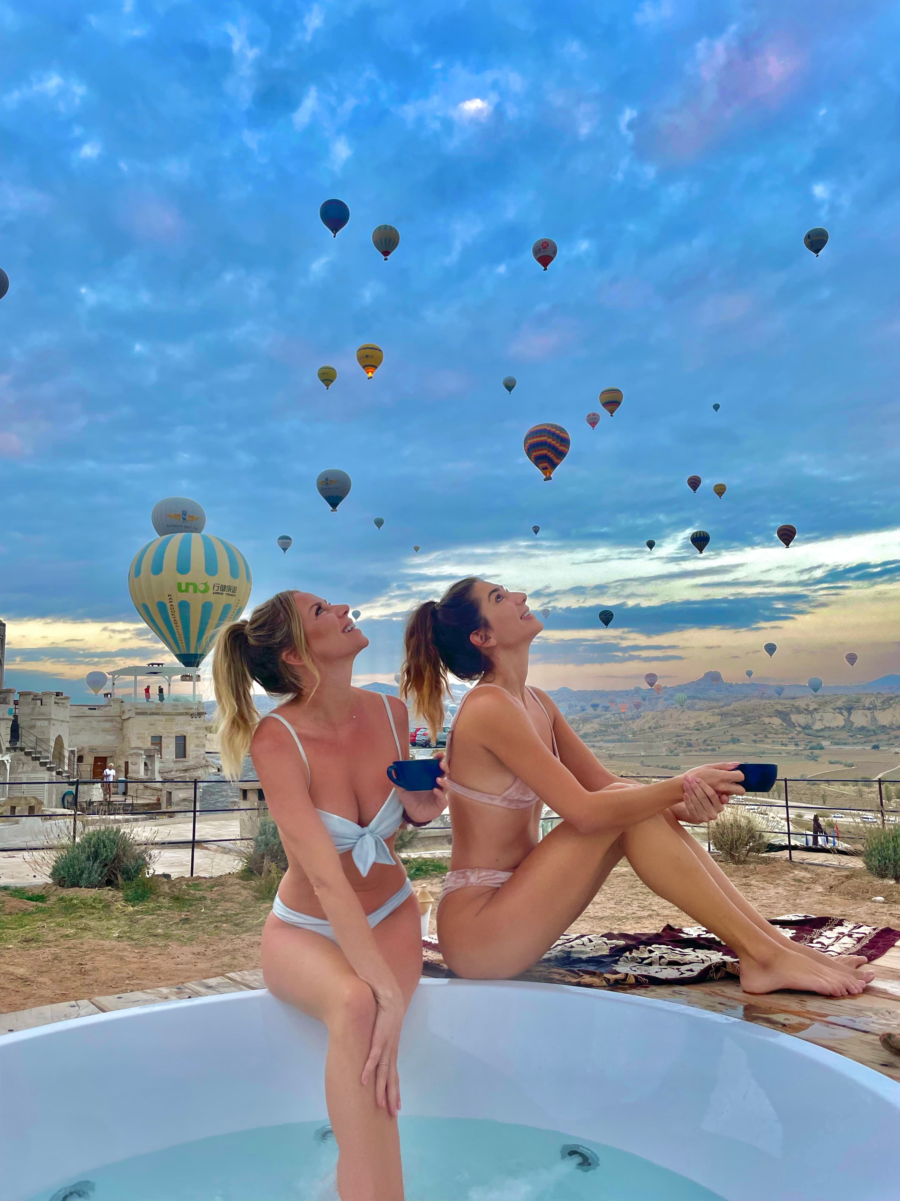 Camille and a friend posing in a jacuzzi overlooking a valley with several hot air balloons in the sky above