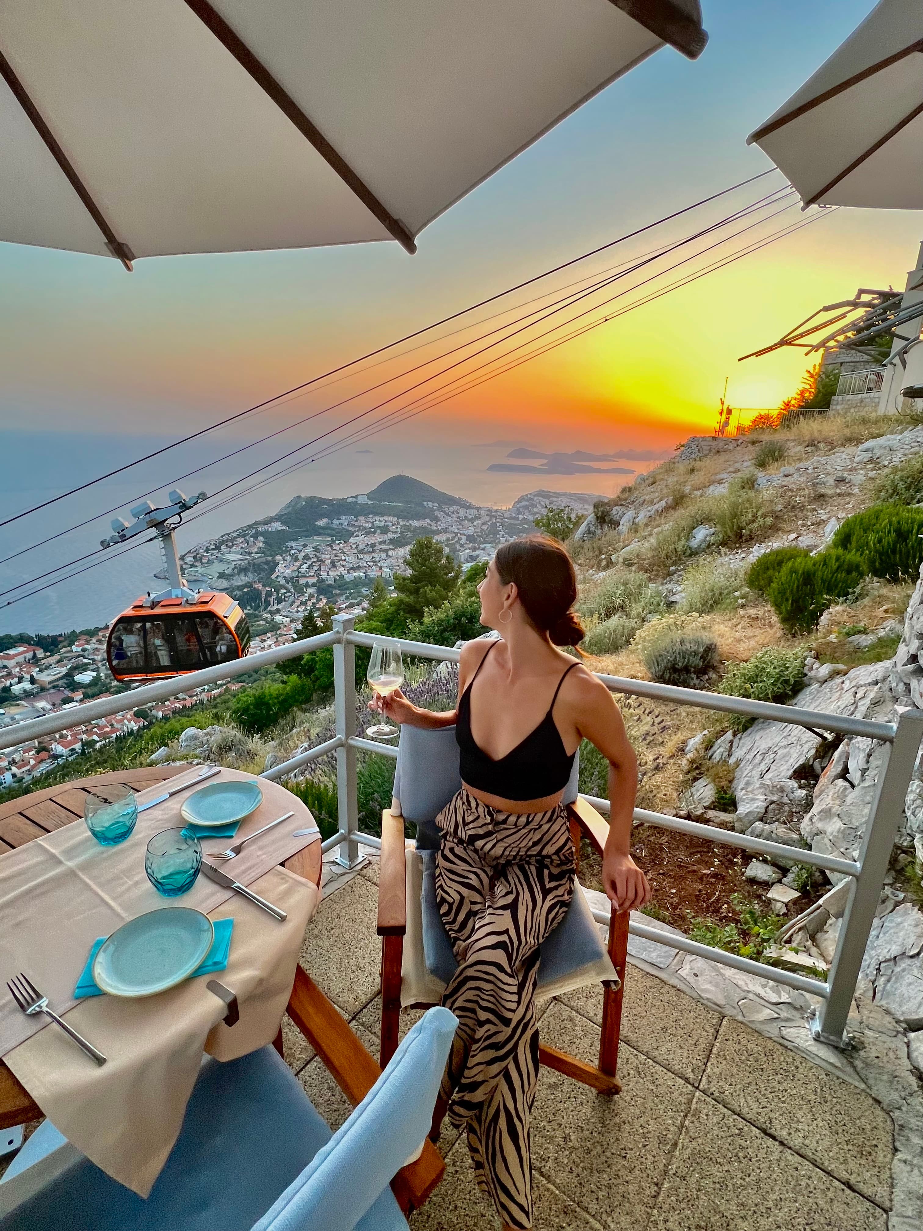Camille seated at a dining table outdoors on a balcony overlooking a coastal area at sunset
