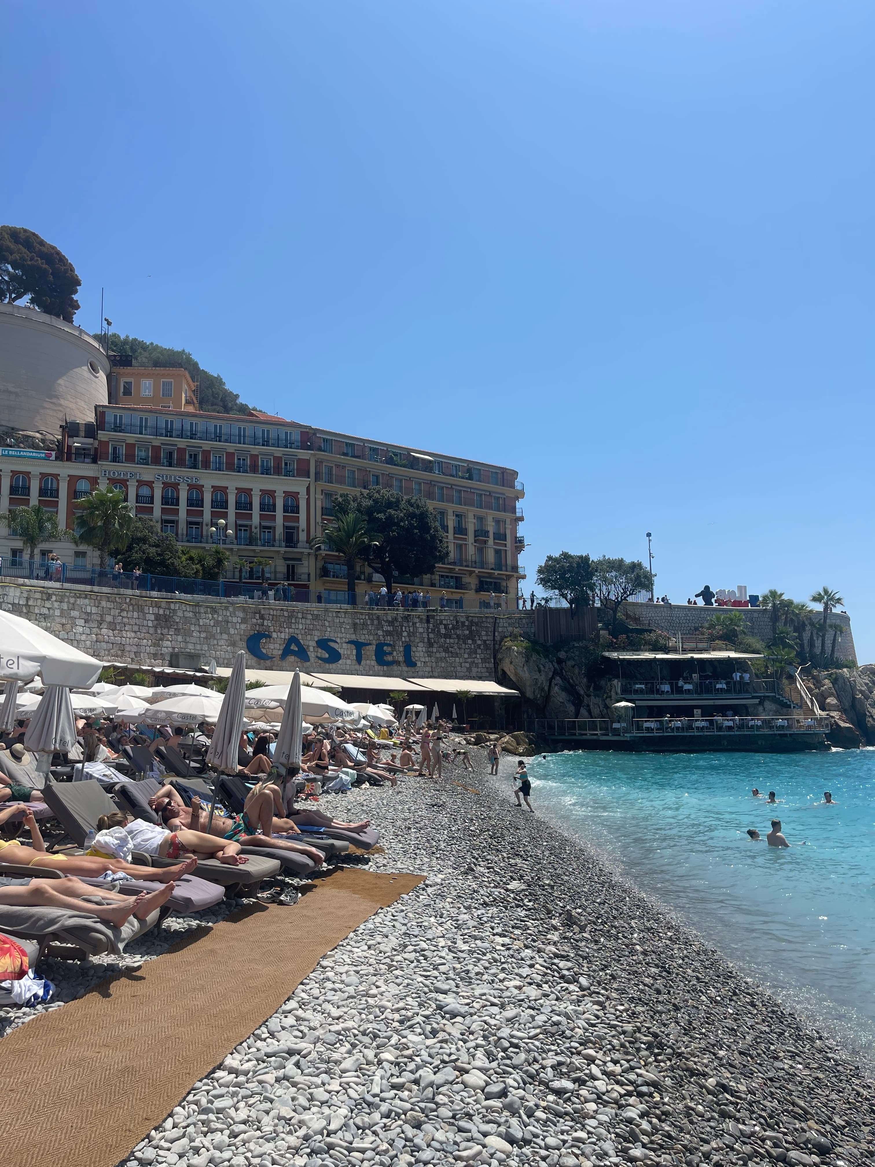 View of a rocky beach and rows of lounge chairs with sunbathers under clear skies