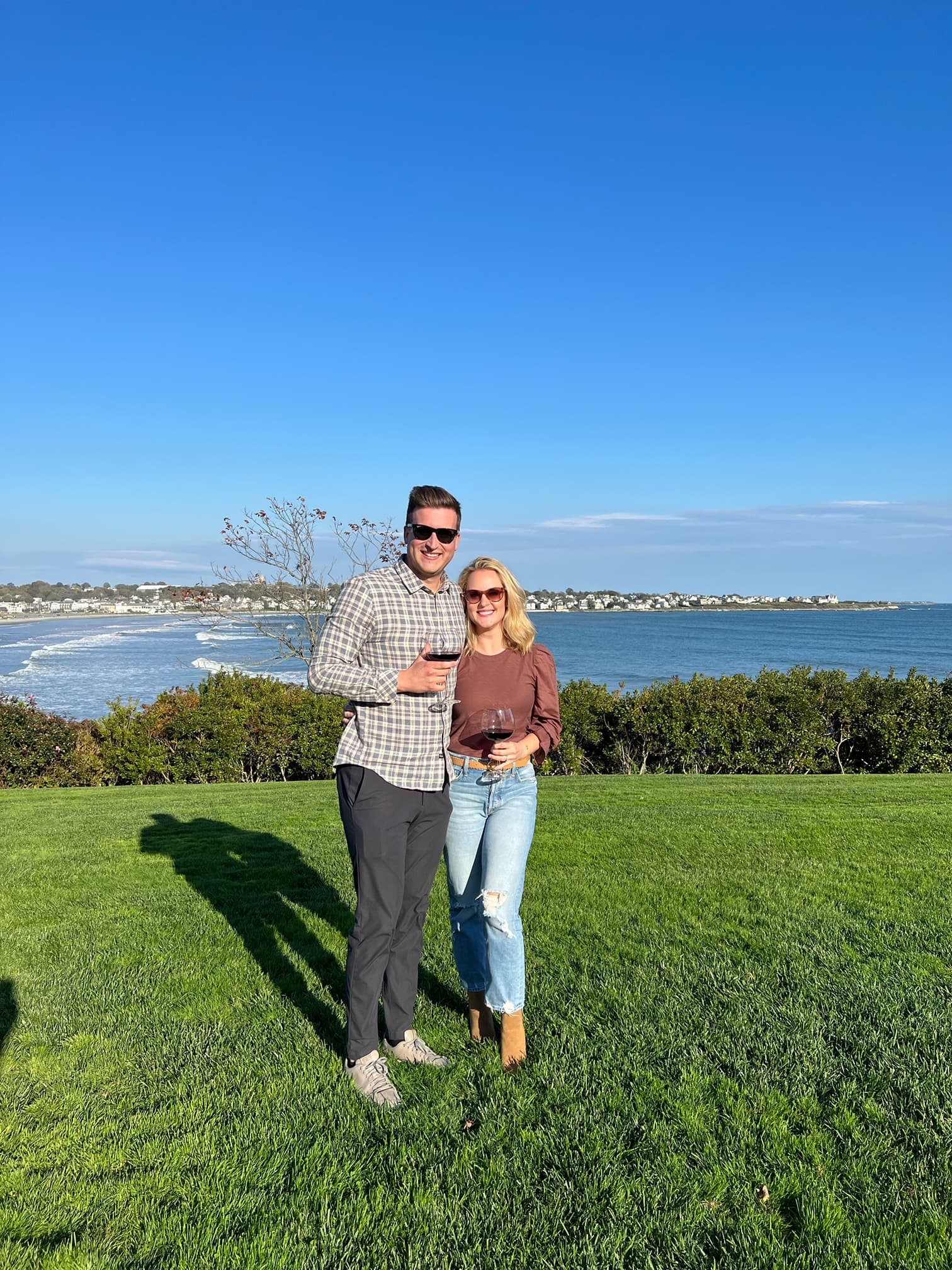 Alex and a man with their arms around each other holding glasses of red wine on a grassy area overlooking the coastline