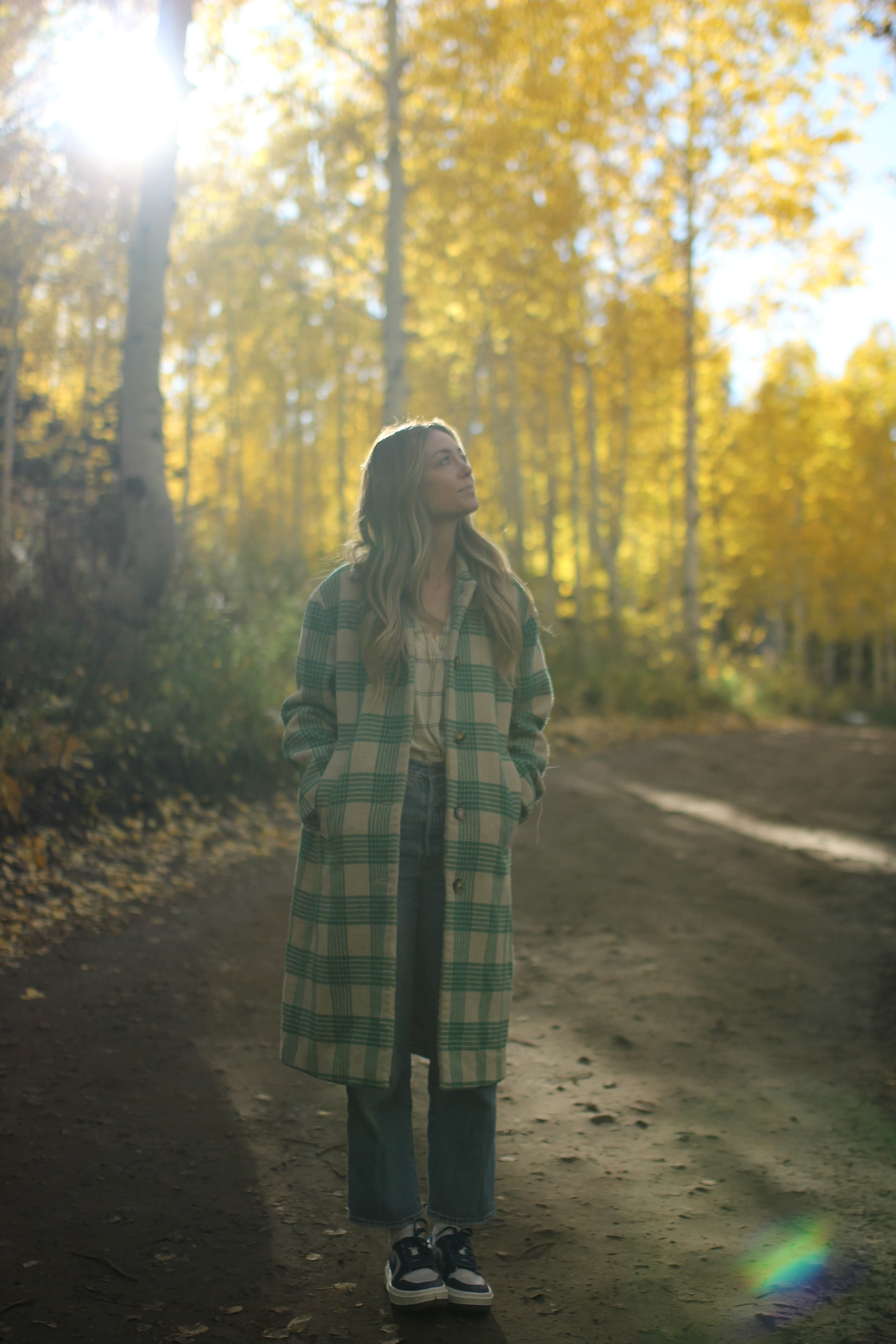 Jill in a long green and white coat with hands in pockets surrounded by yellow trees