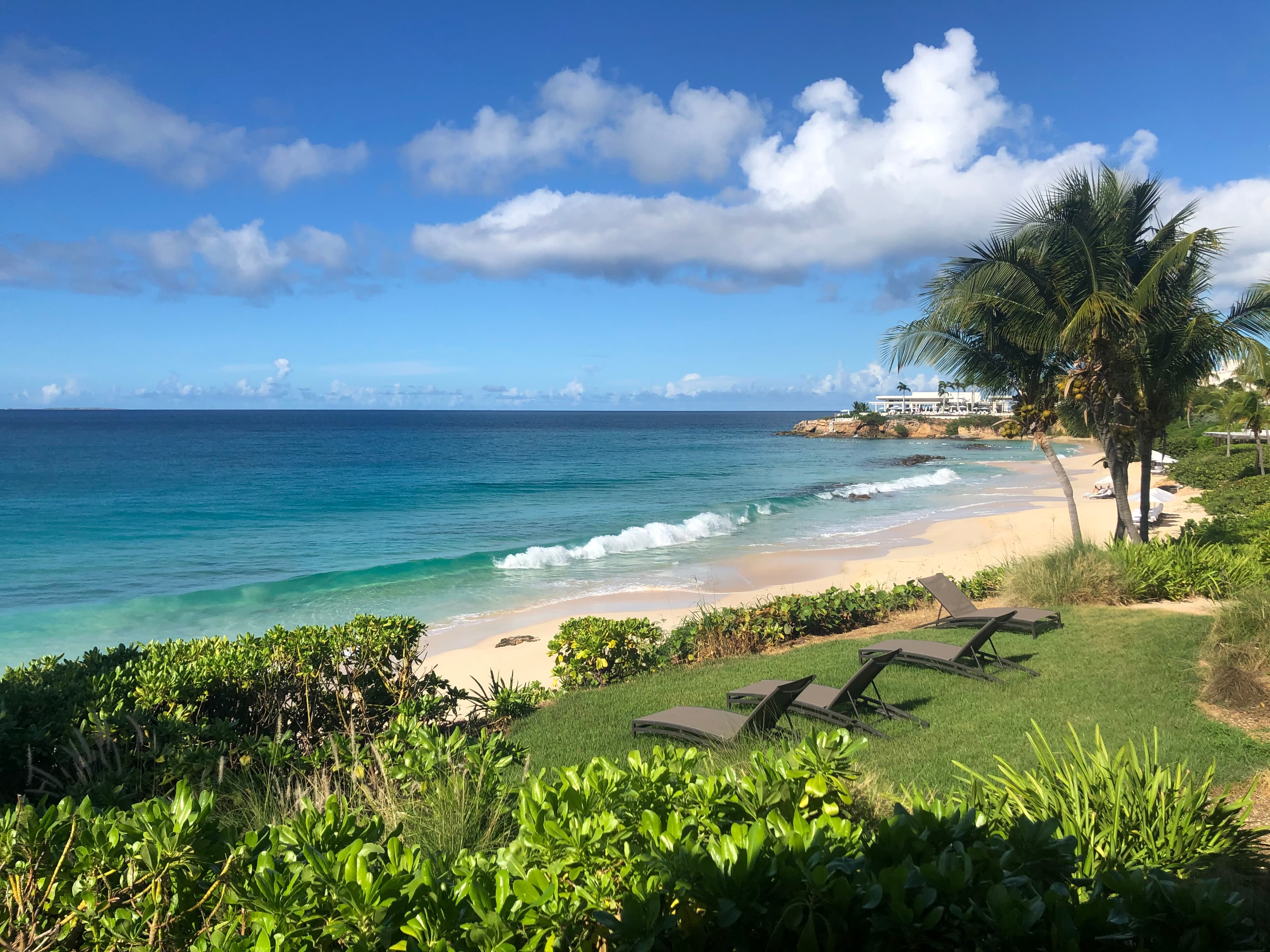 Beautiful view of four lounge chairs looking out over an empty white sand beach and calm waves on a sunny day