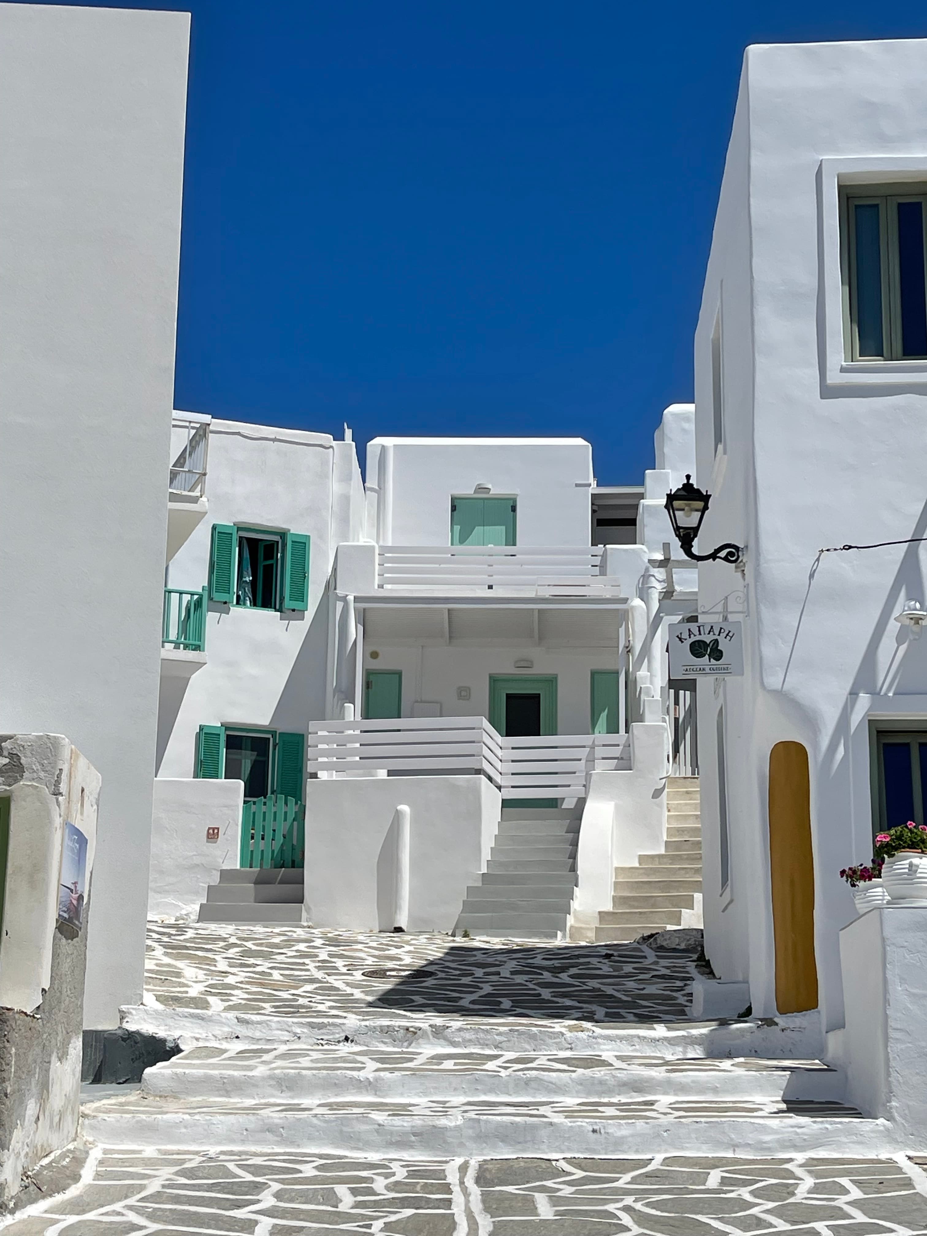 White buildings with green shutters and balconies line a narrow, stone-paved street under a clear blue sky.