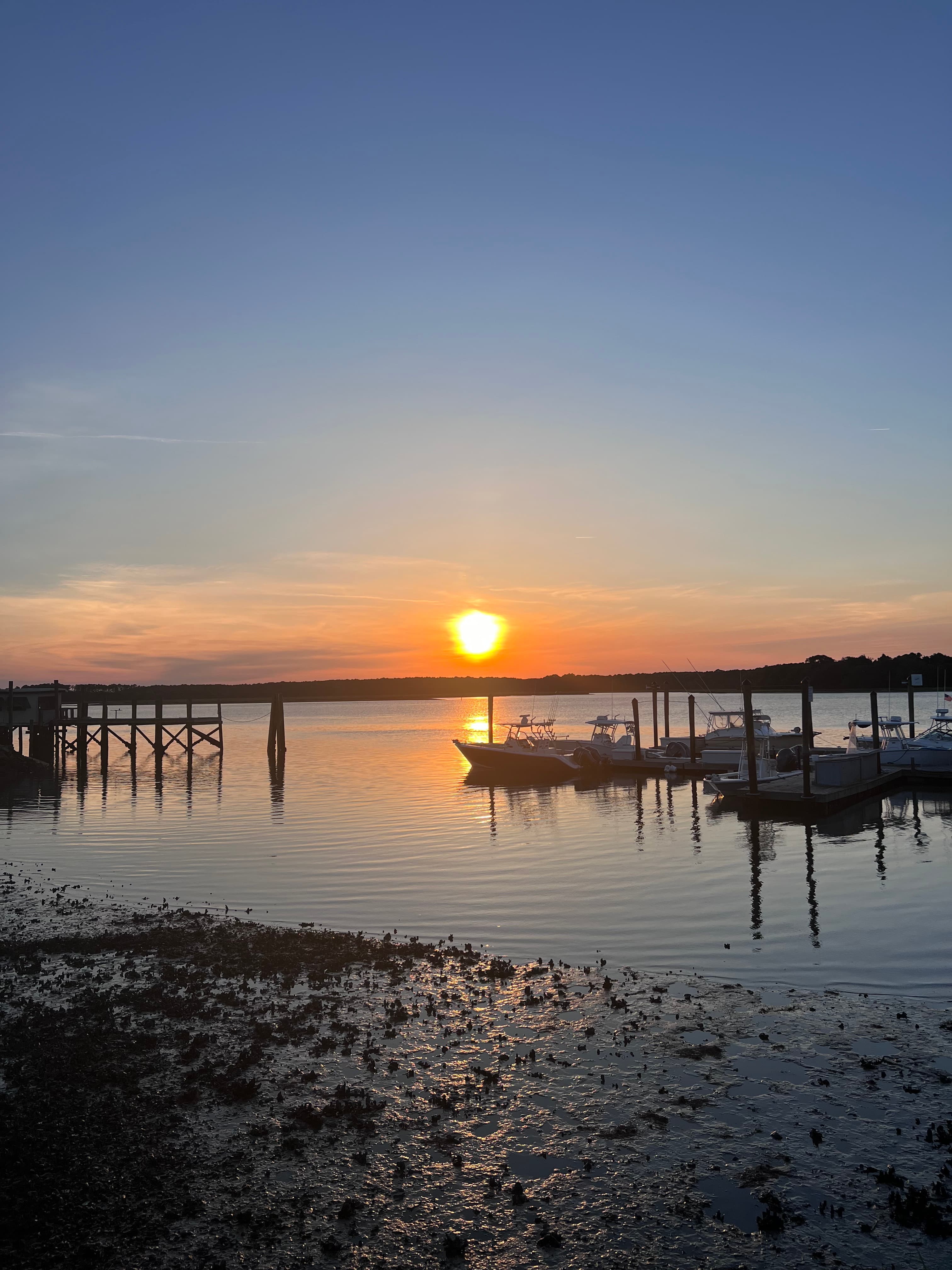 The sun sets over still water near the shore as boats float across the pier on a clear day.
