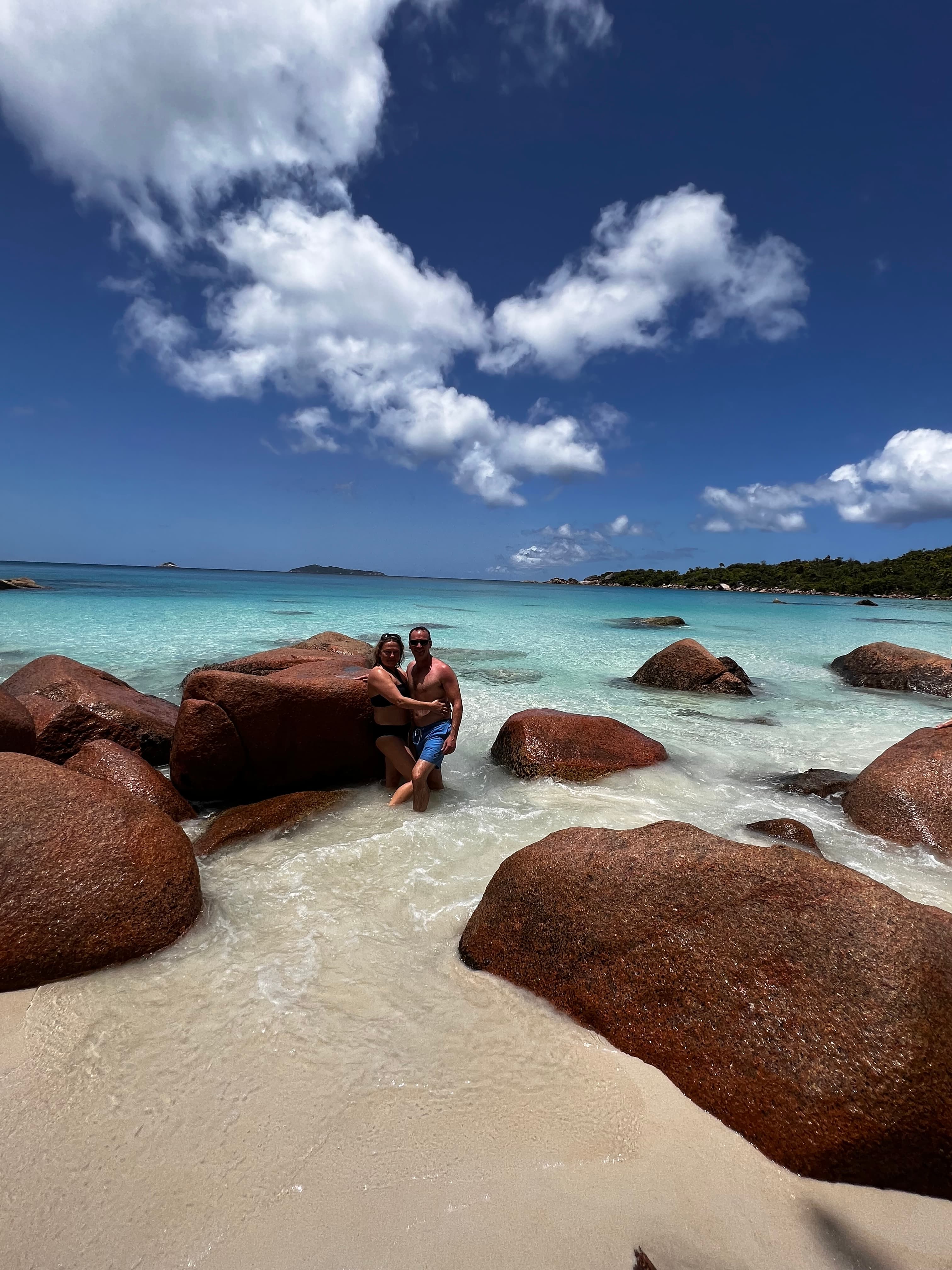 Large clay colored rocks lay on the white sand beach as waves gently lap the shore on a clear day.