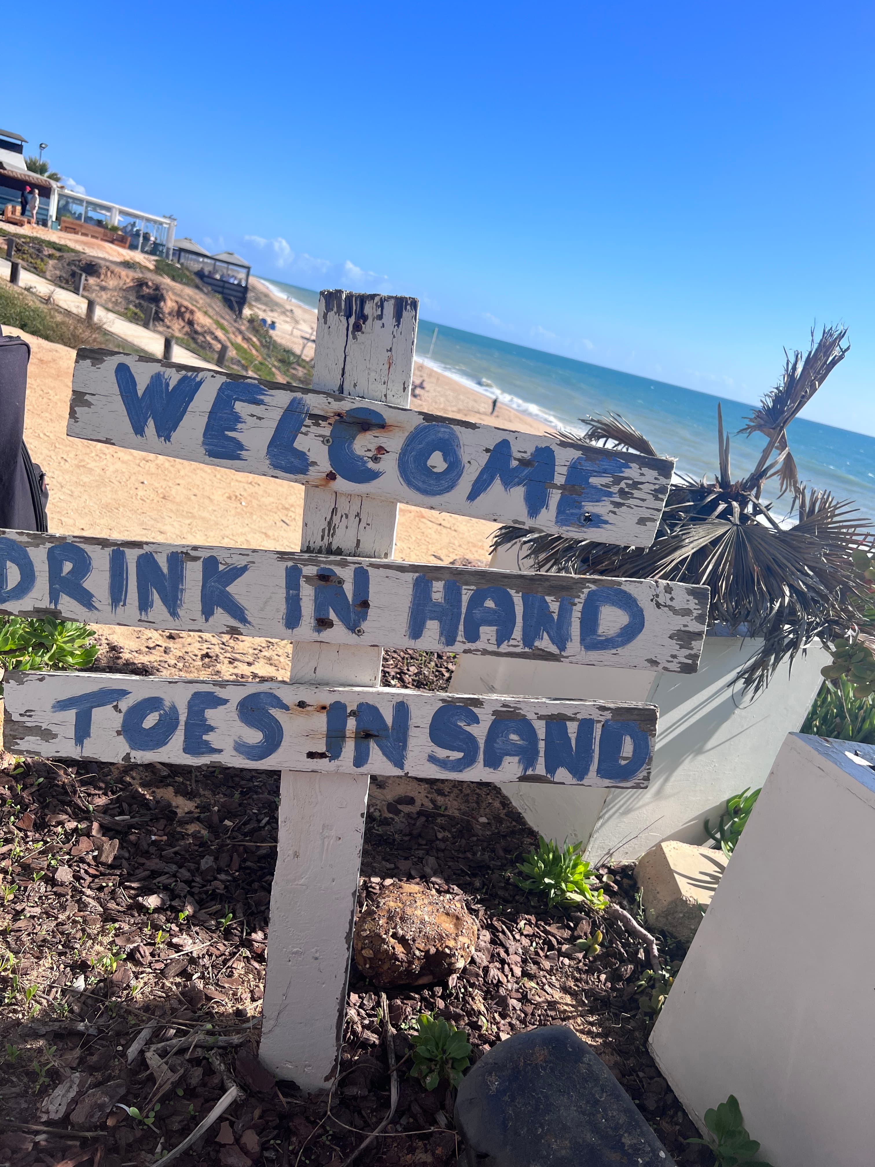 Beach sign posted before the shore reading "drink in hand, toes in sand" on a sunny day.