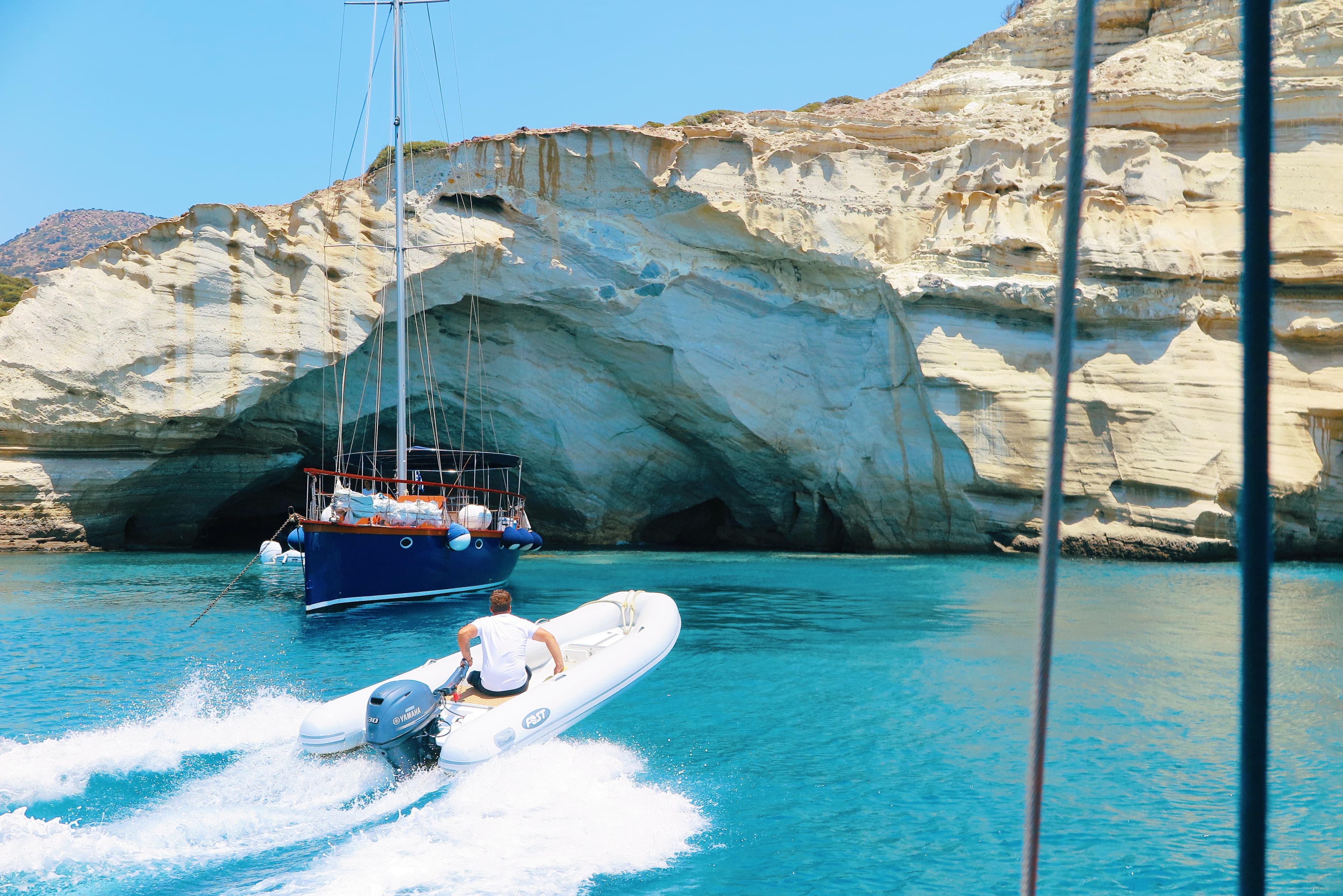 A person rides a motorboat towards a rocky cliffside with a cave opening, while a sailboat is anchored nearby in the clear blue waters.