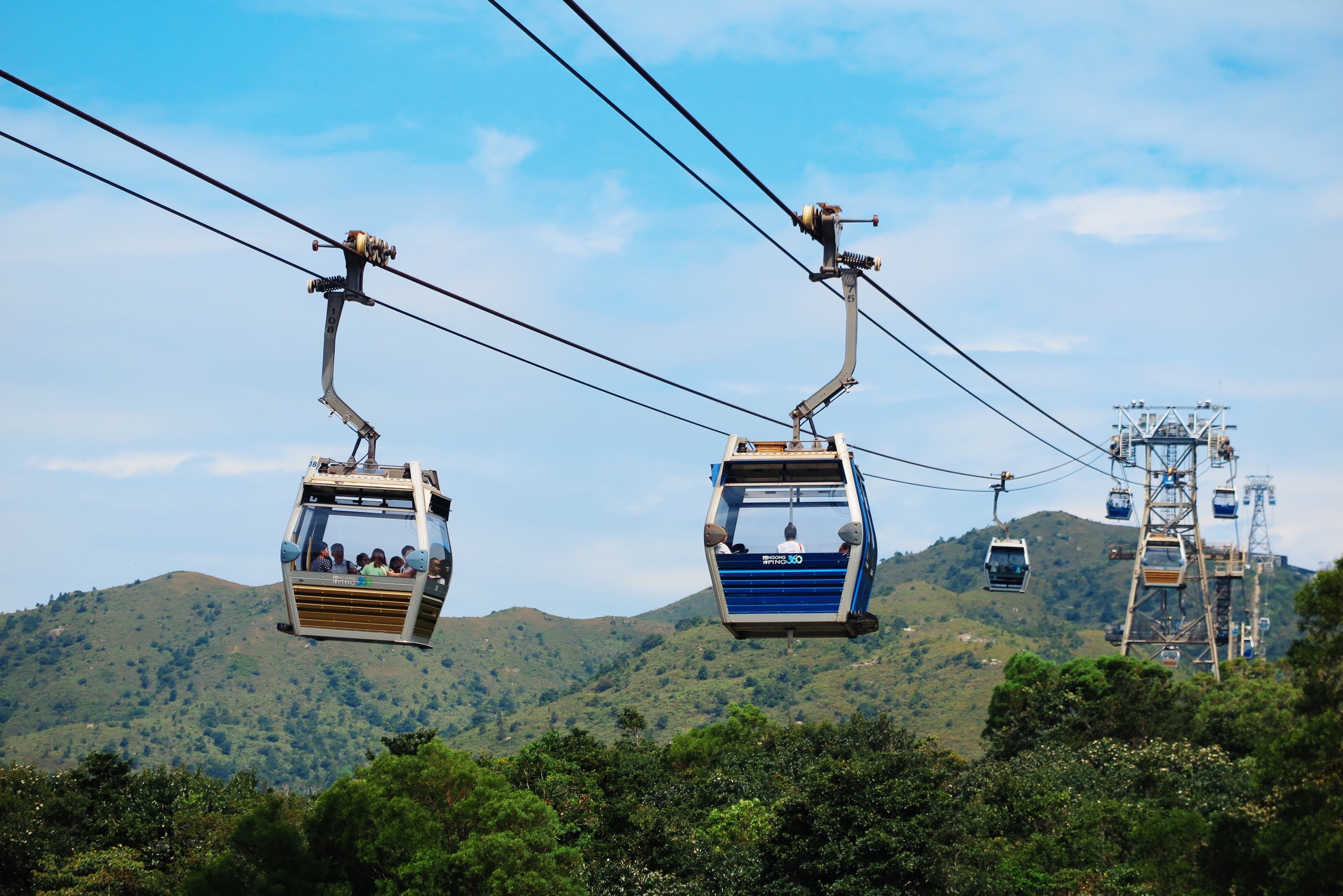 Several cable cars travel along wires above a green, mountainous landscape under a blue sky with scattered clouds.