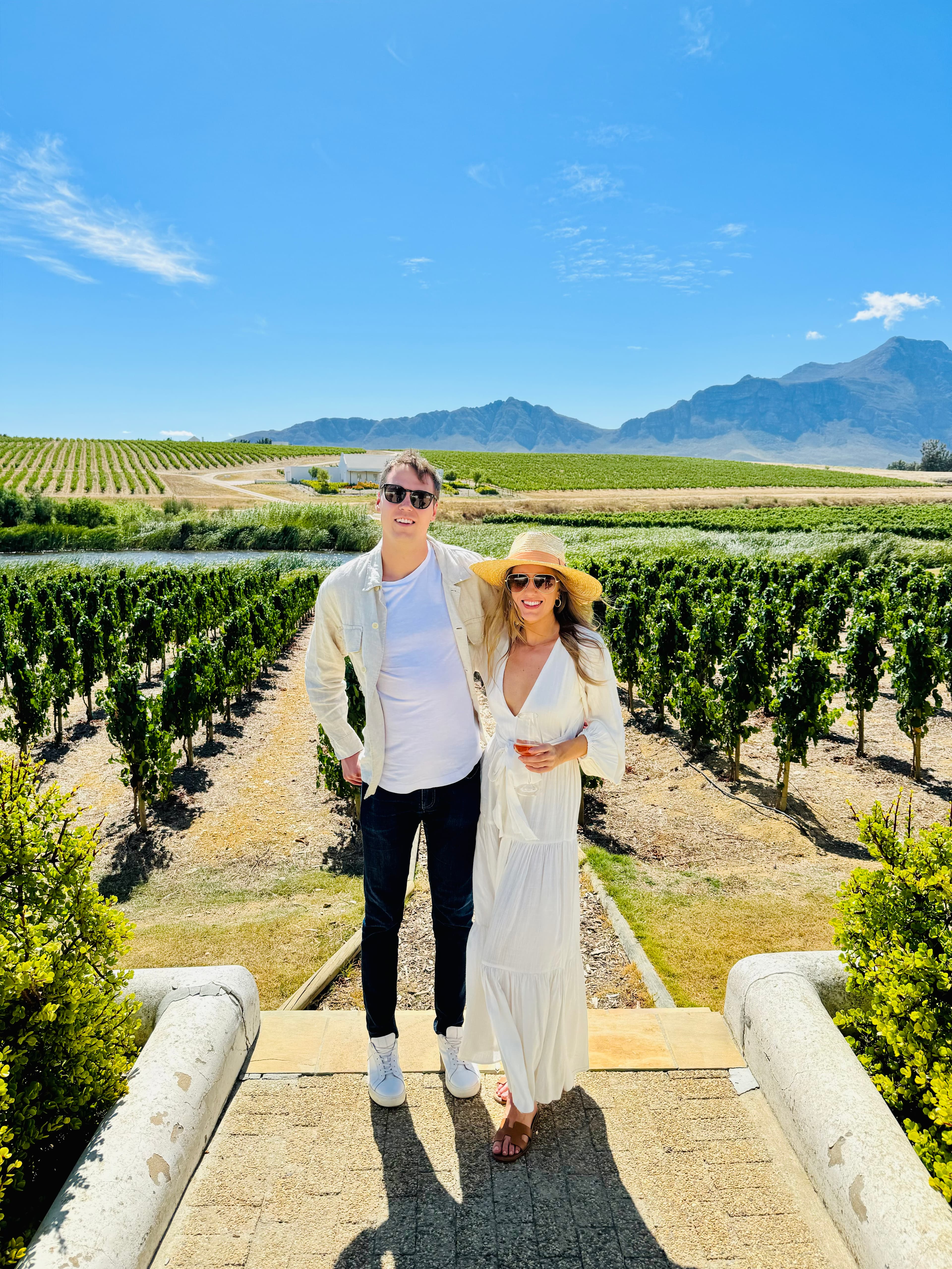 Advisor and her partner in white clothing posing by a vineyard with mountains visible in the distance