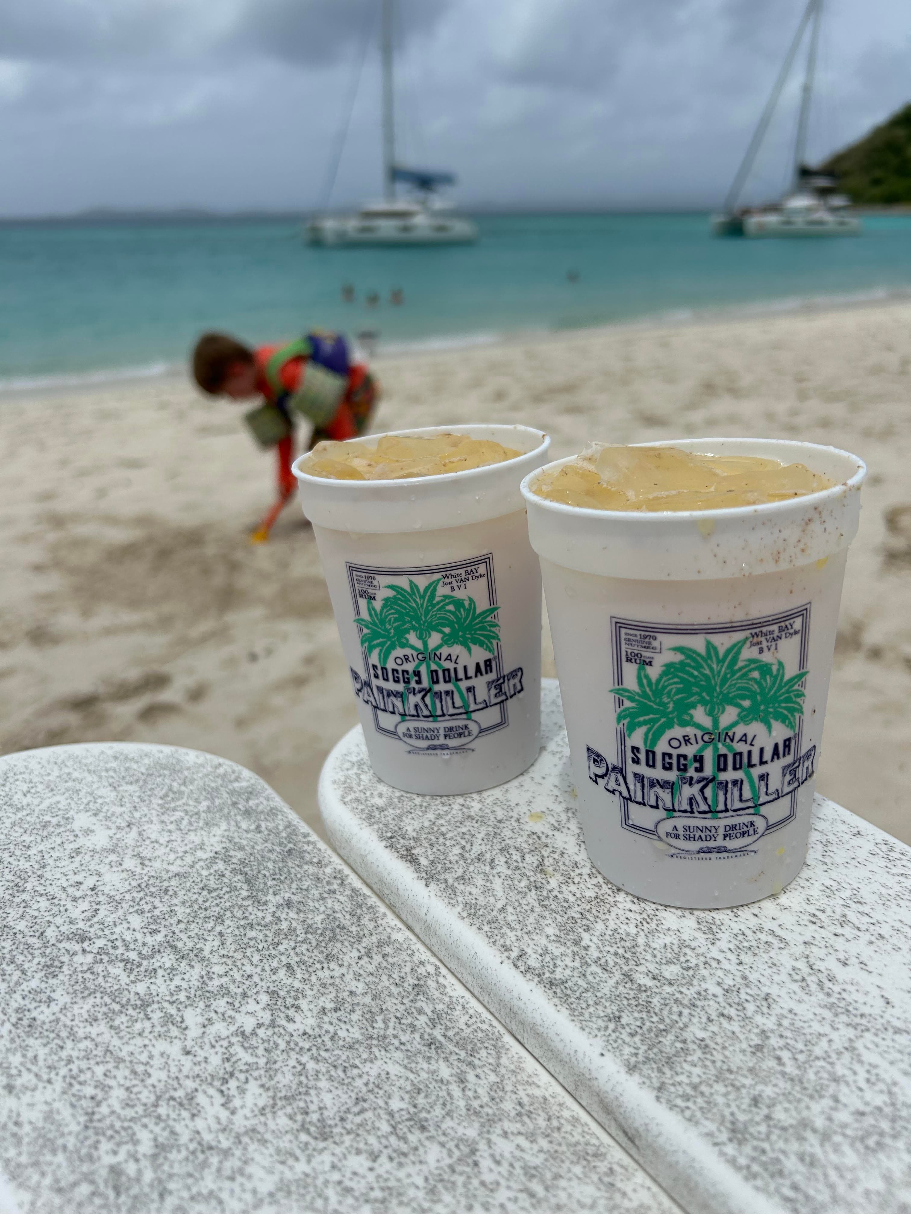 Two cups with drinks inside sitting on a chair on the beach, with the ocean in the background.