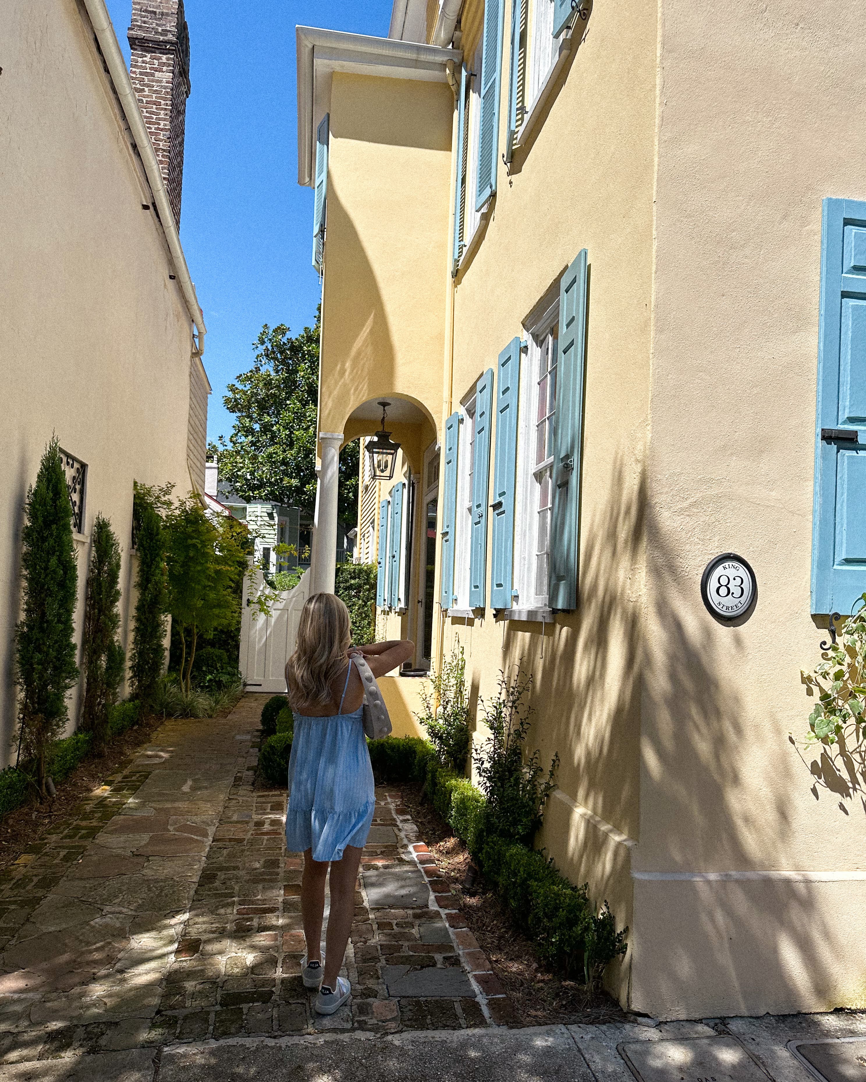 A woman wearing a blue dress and looking out to a view of a house with blue windows and a cream colored exterior.