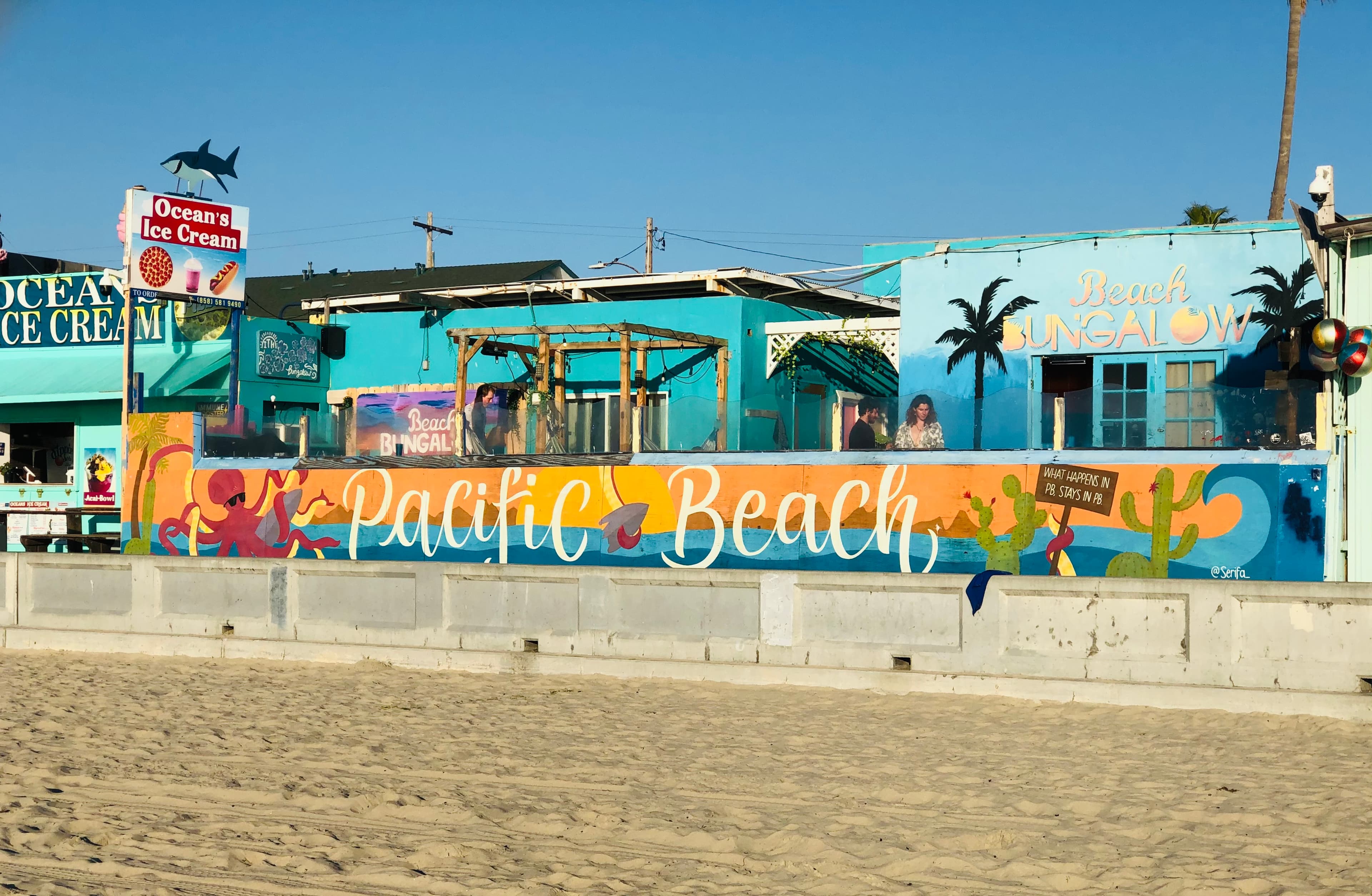 View of a colorfully painted beachside mural reading “PACIFIC BEACH” with blue buildings behind it