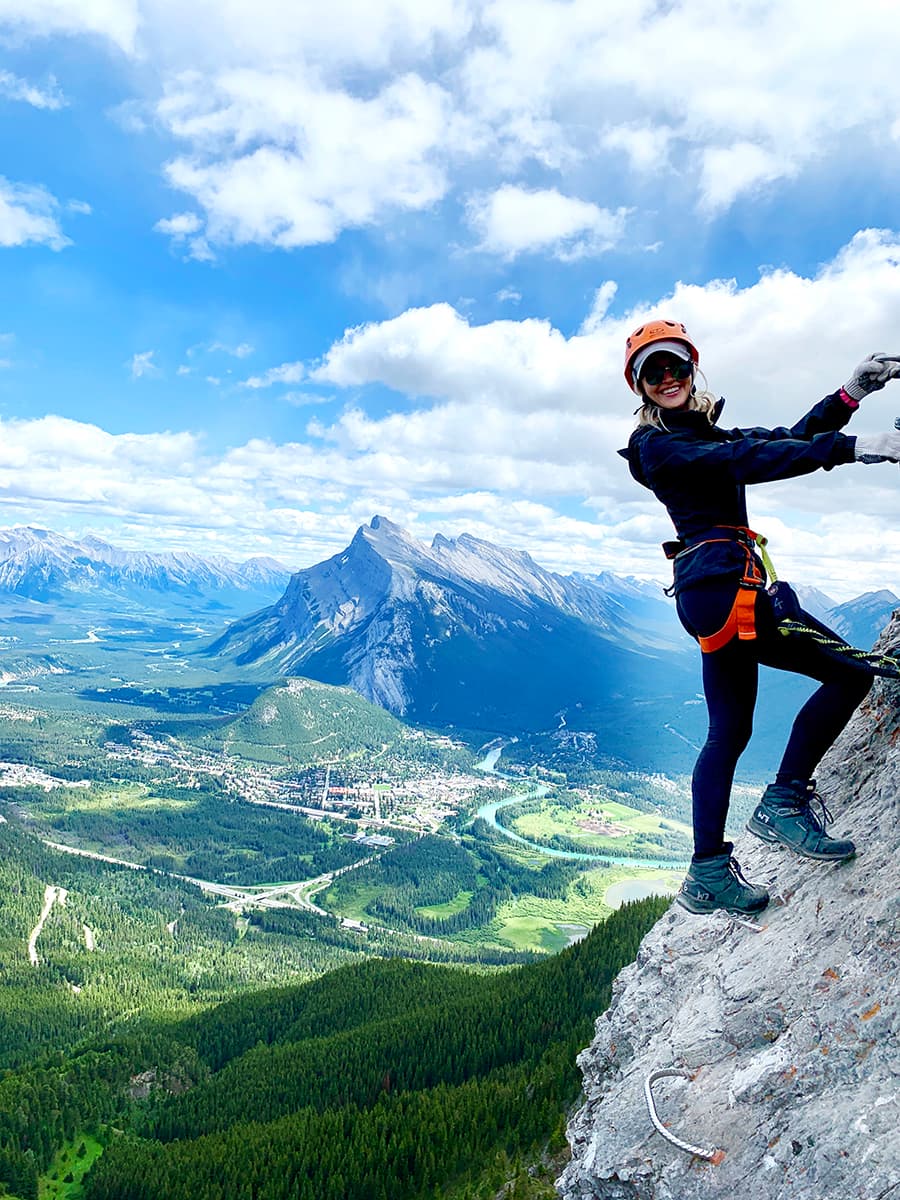 Advisor in rock climbing gear seen on the side of a mountain with a beautiful green valley behind her