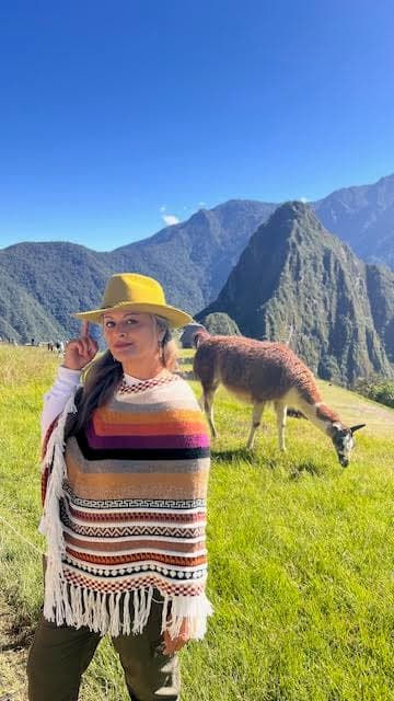 Advisor posing besides an alpaca on a grassy plain with mountains in the background