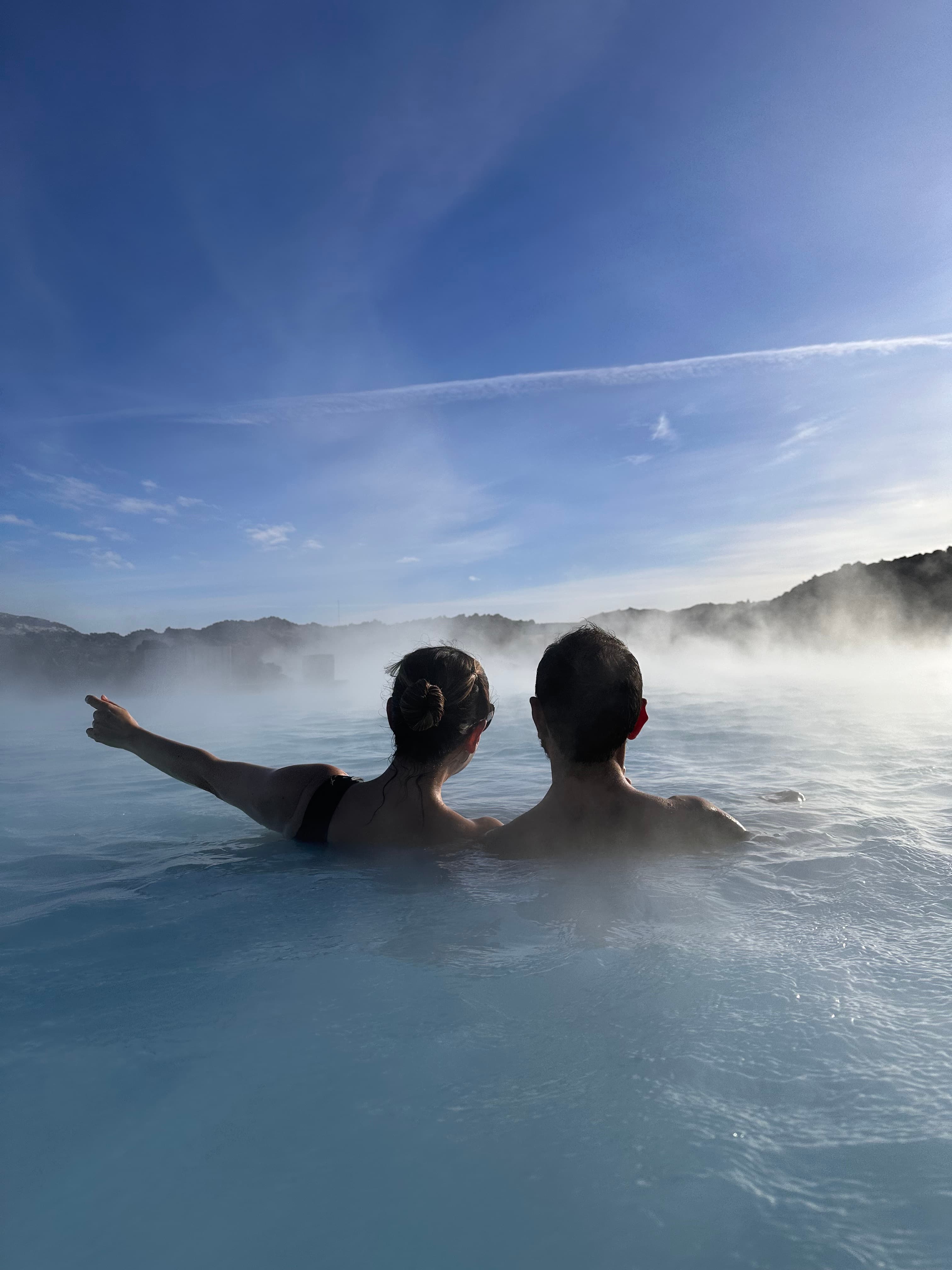 Karla and partner in the water of a geothermal pool. They are viewed from behind, with one person pointing towards something in the distance.