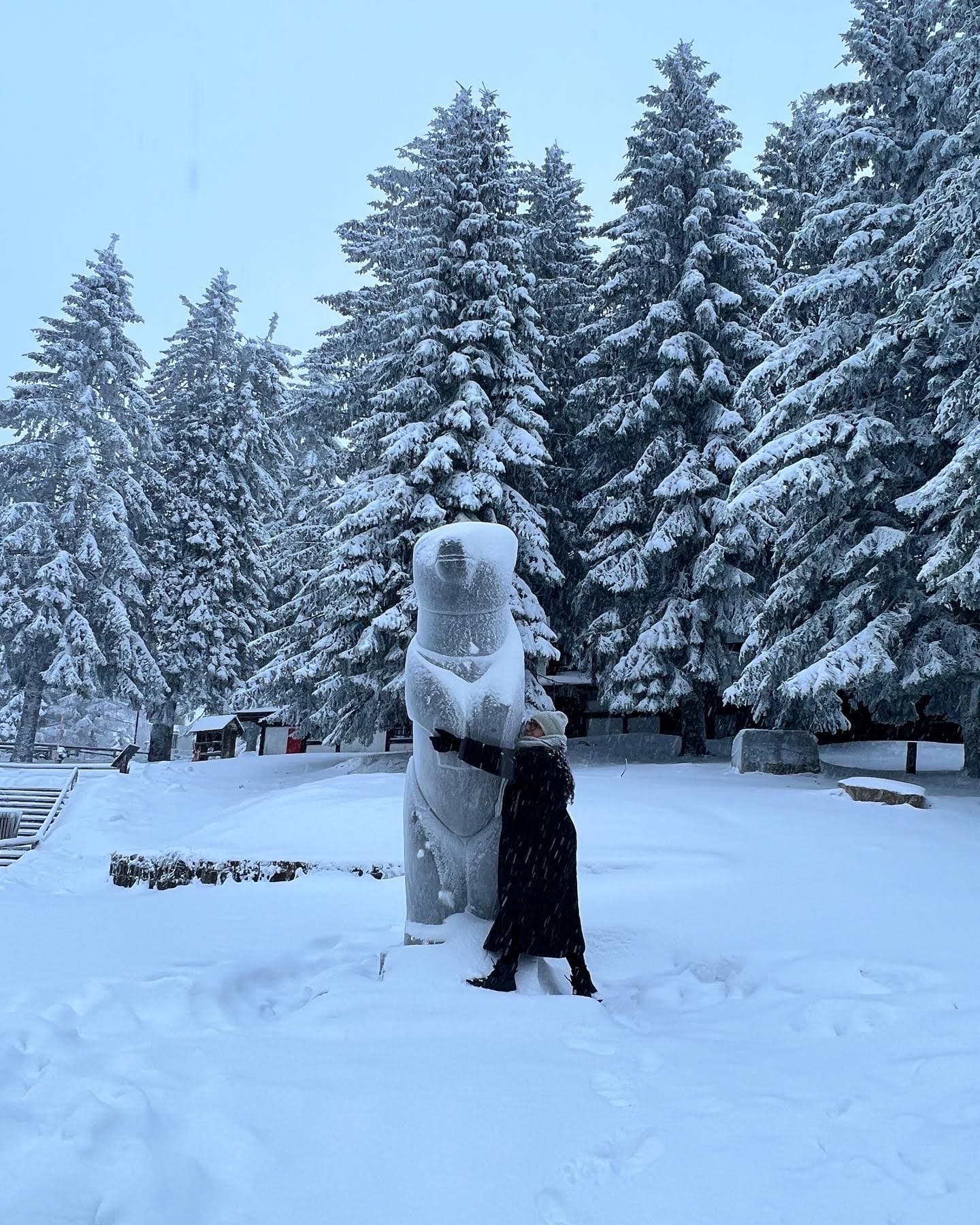 View of a person in a long black coat posing in front of a polar bear statue in a snowy environment