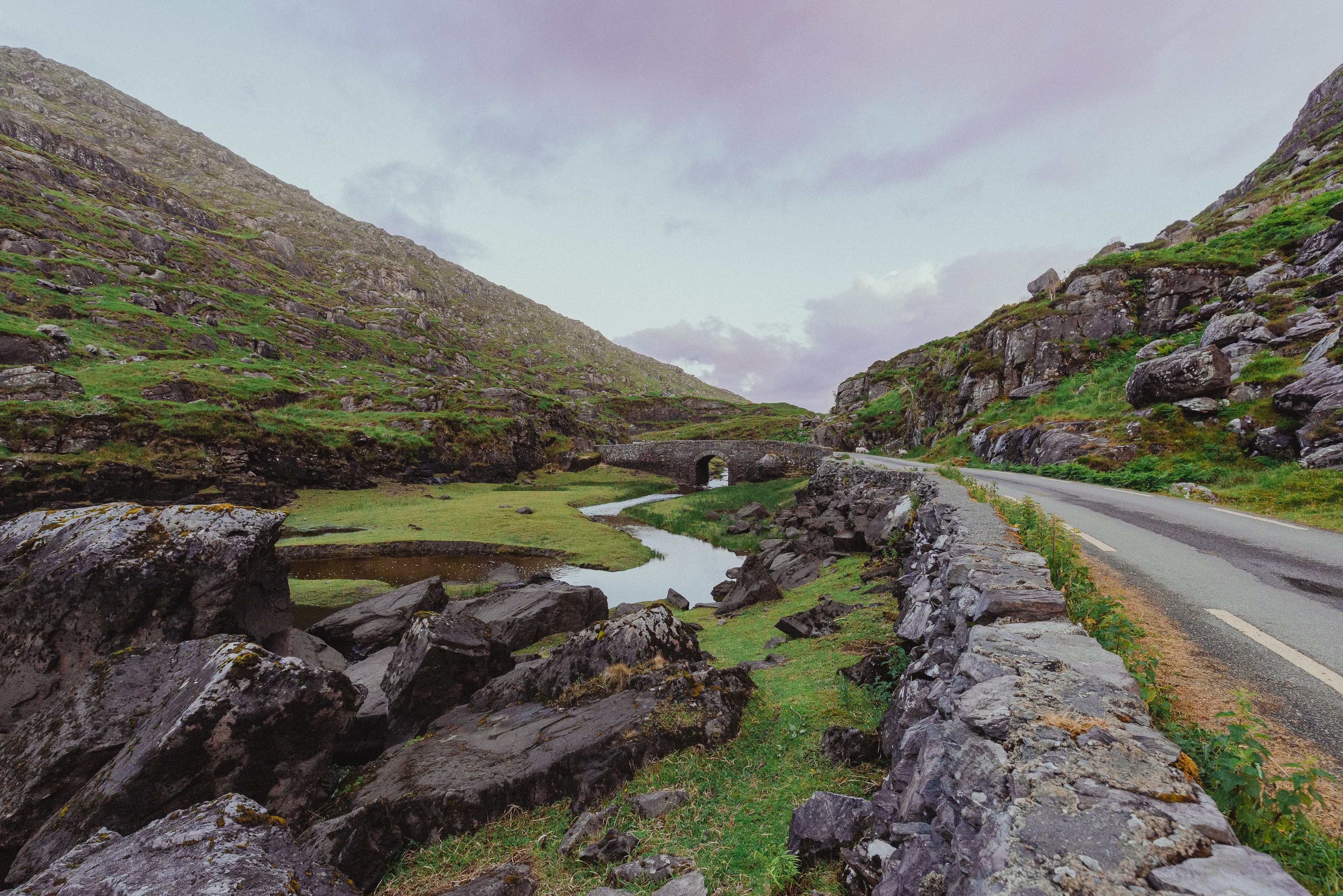View of an empty road running through a green valley under cloudy skies