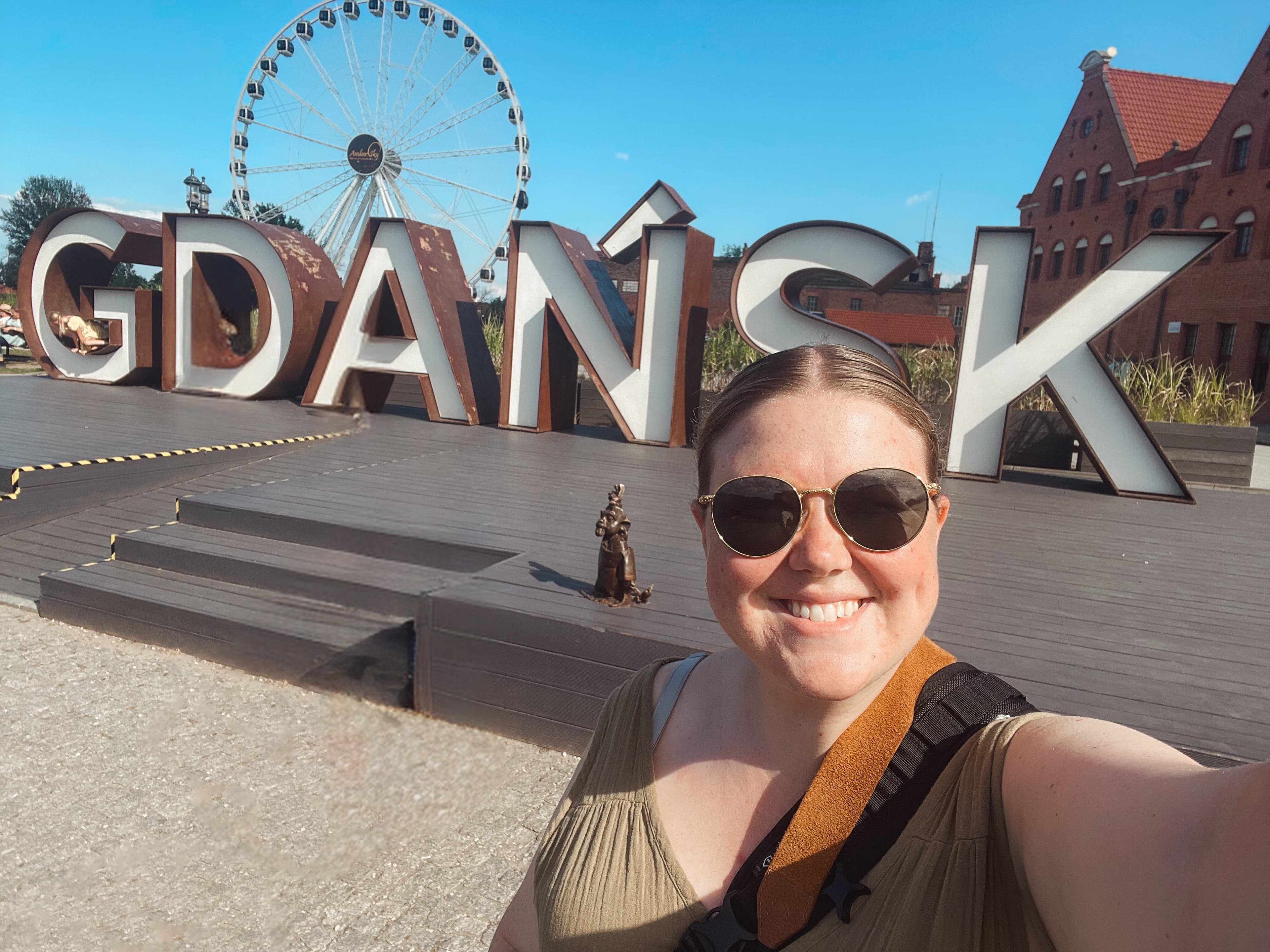 Emily taking a selfie in front a large sign of letters spelling “GDAŃSK” with a Ferris wheel in the background