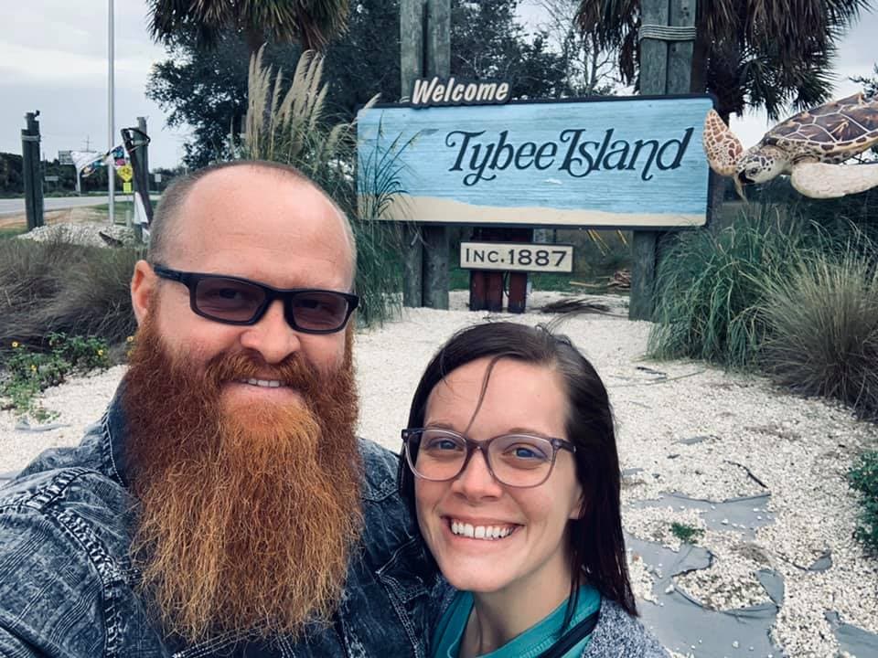 Advisor and a man posing in front of a blue sign that reads "Tybee Island, inc. 1887" on a sandy beach.