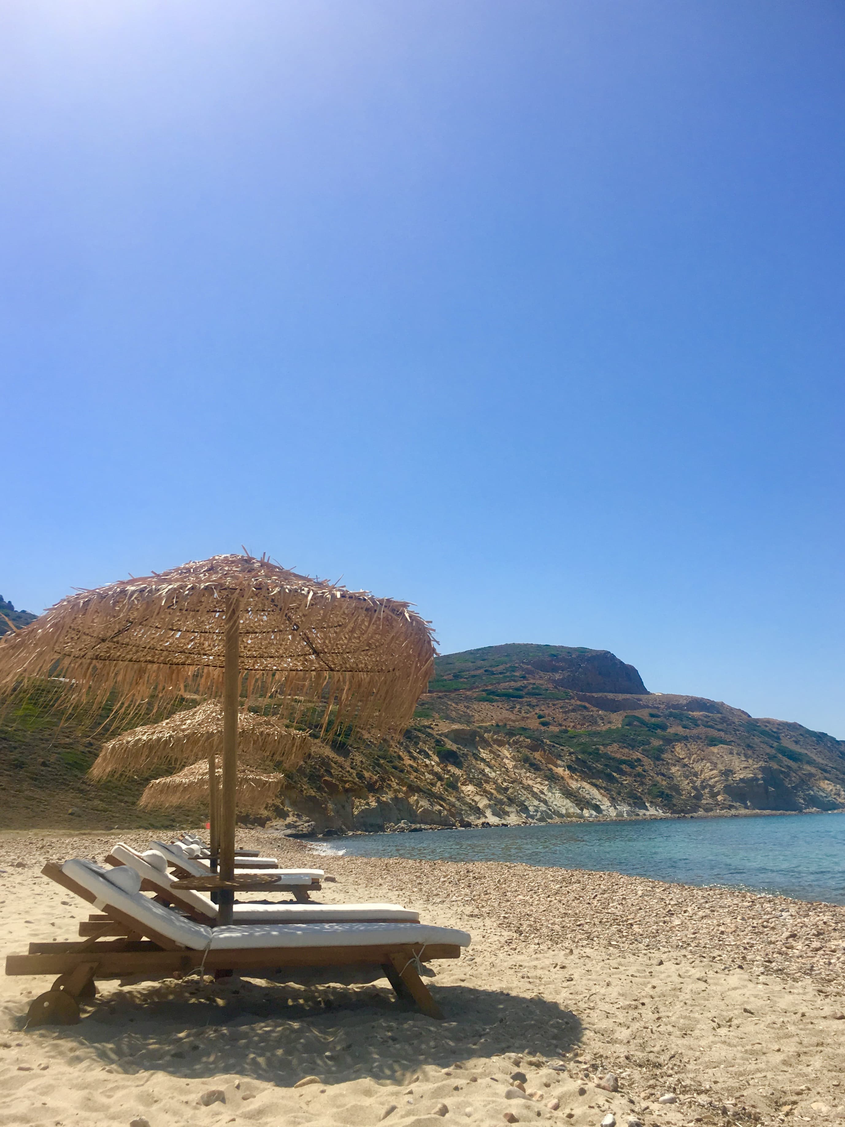 Loungers and beach umbrellas line the shore of a sandy beach as waves slowly roll in on a sunny day.