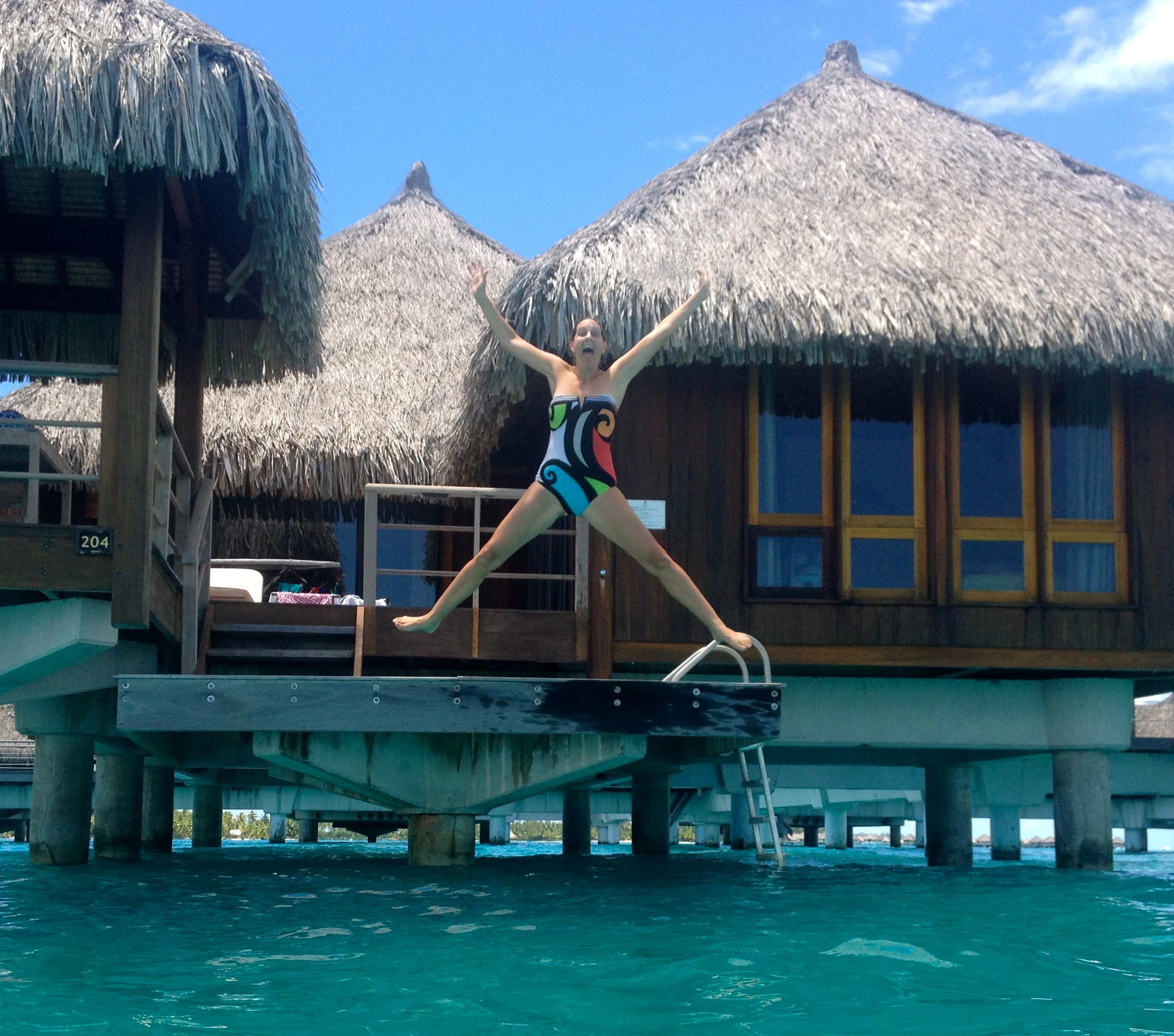 Veronica jumping into the ocean, with huts behind her.