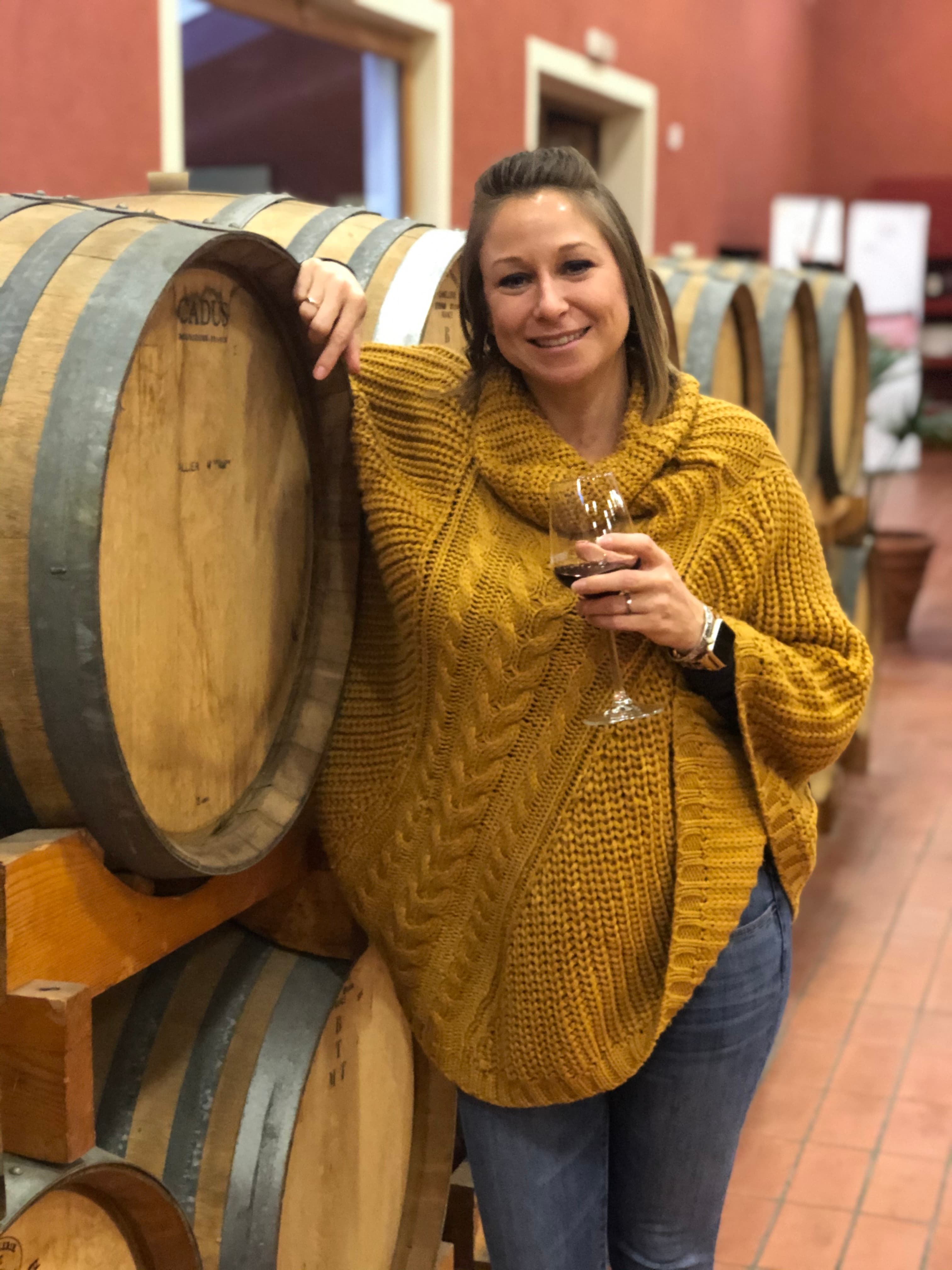 A woman posing next to a wooden barrel while holding a glass of wine and wearing a yellow knit sweater.