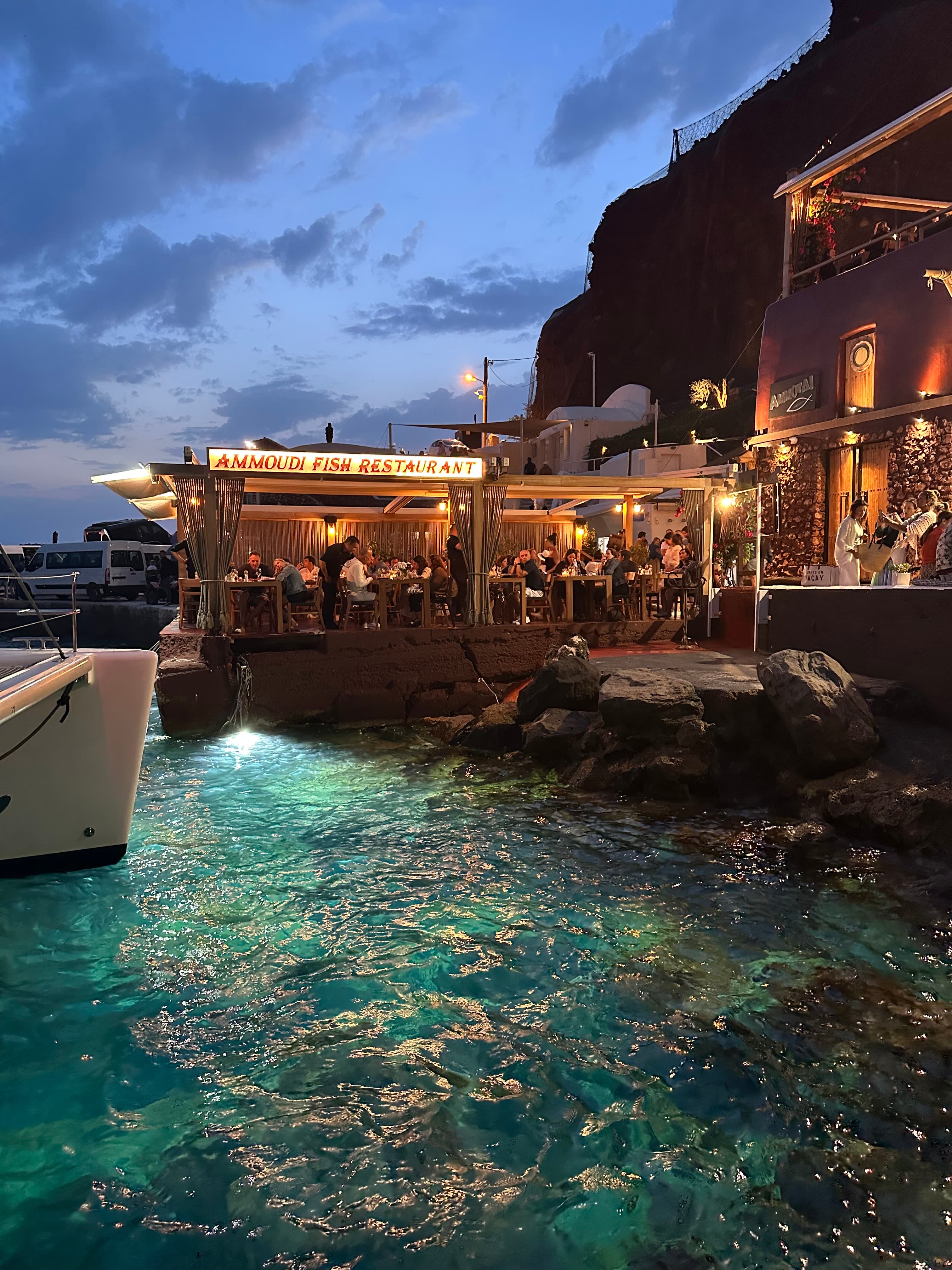 A restaurant patio lit up near the water on a night dotted with clouds.