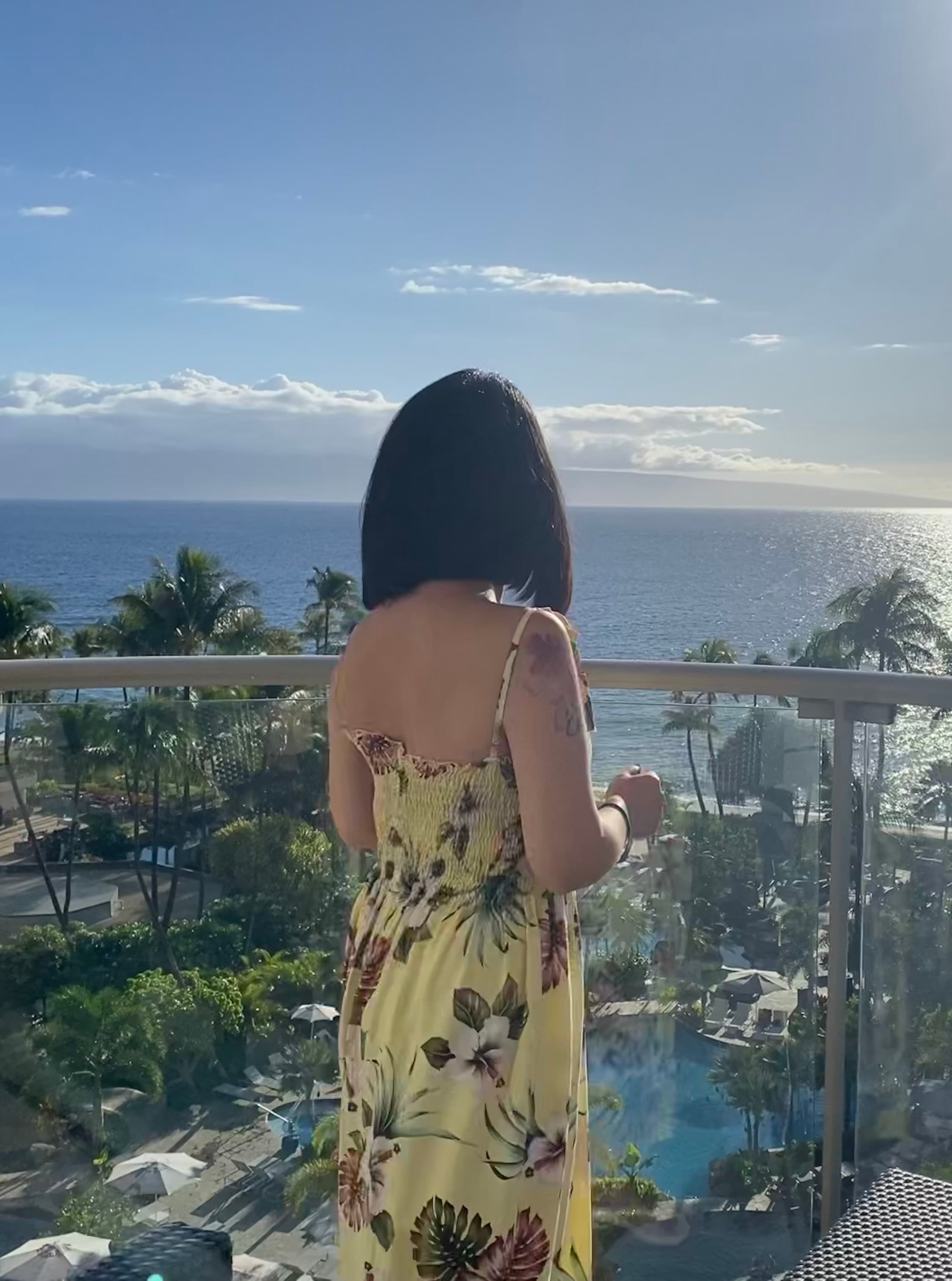 Advisor in a yellow floral print dress standing on a glass balcony overlooking a resort and the sea