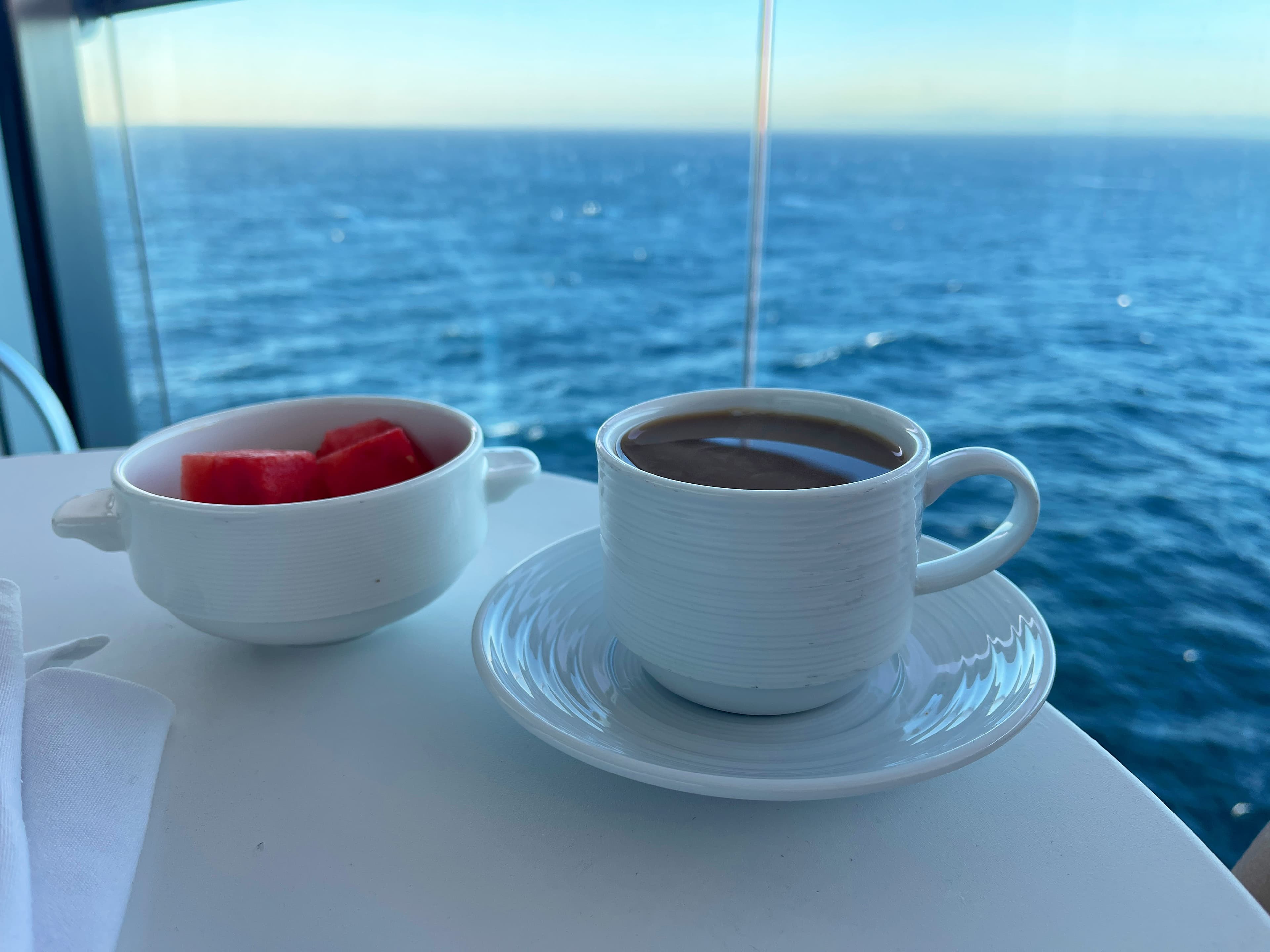 A coffee mug and bowl of fruit on top of a white table next to a window that looks out to the blue sea.