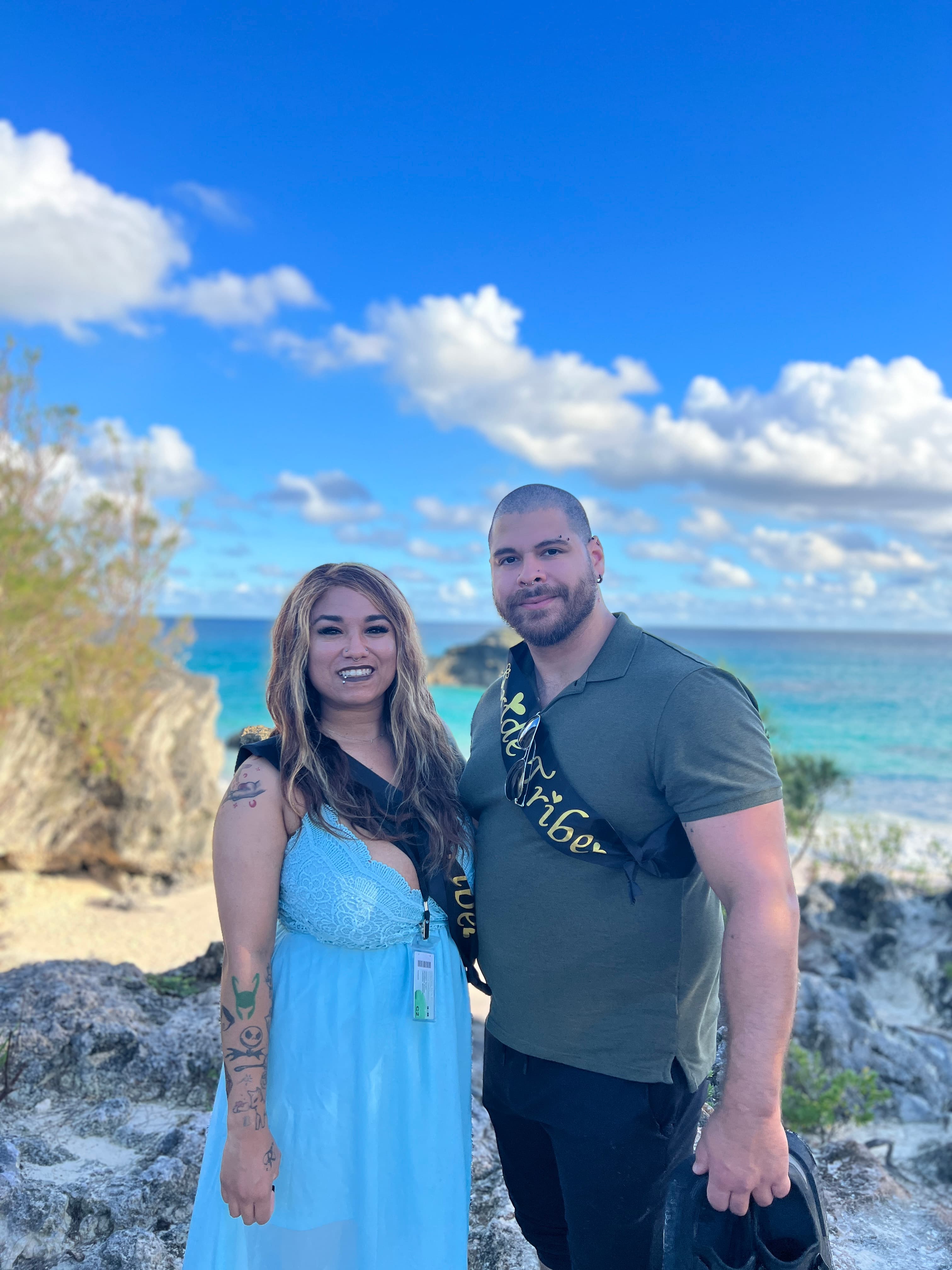 A couple posing in front of a body of water and a beautiful day.