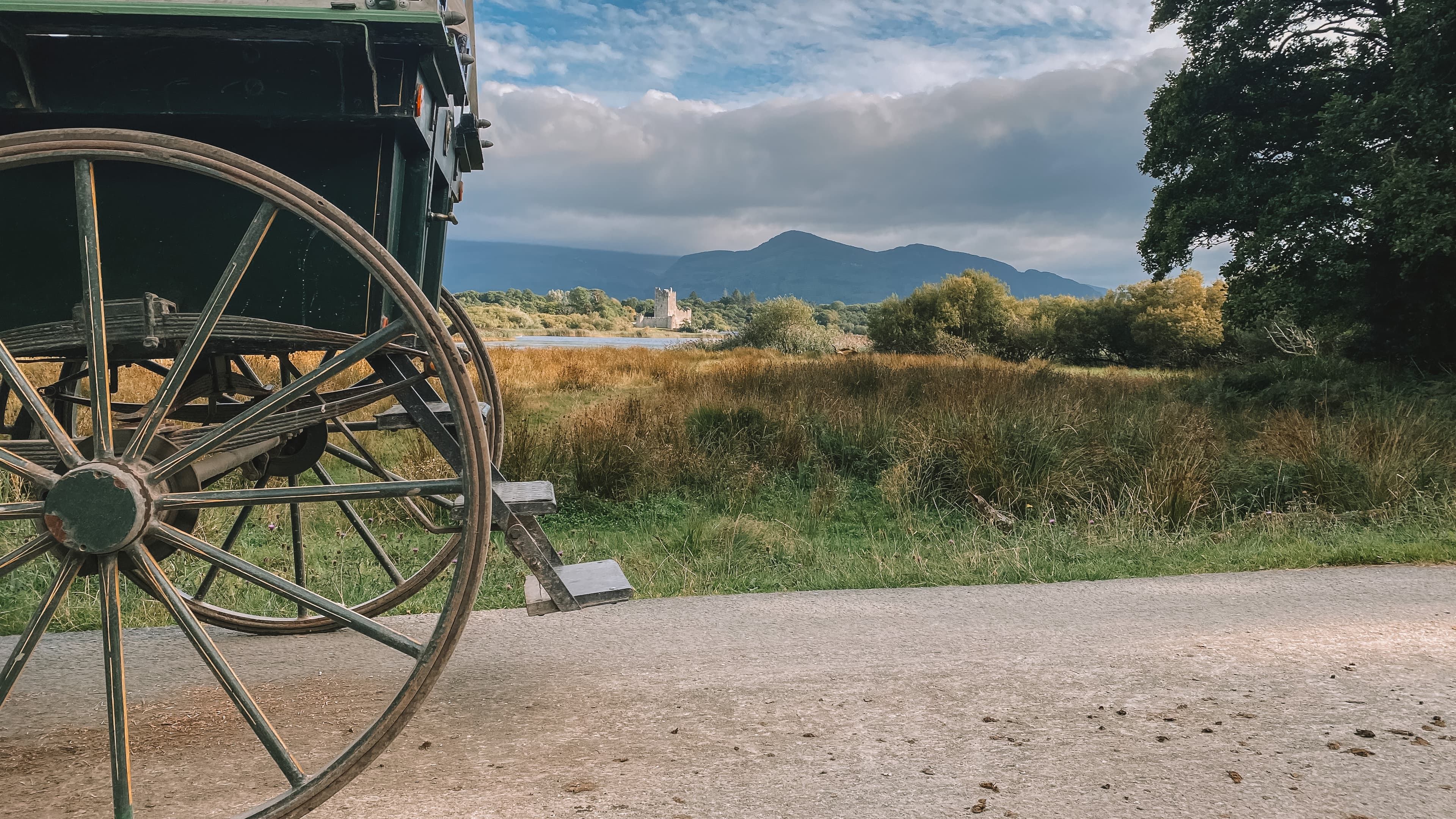 A carriage and wagon wheel sit on a dirt path under a cloudy sky as mountains frame the horizon.