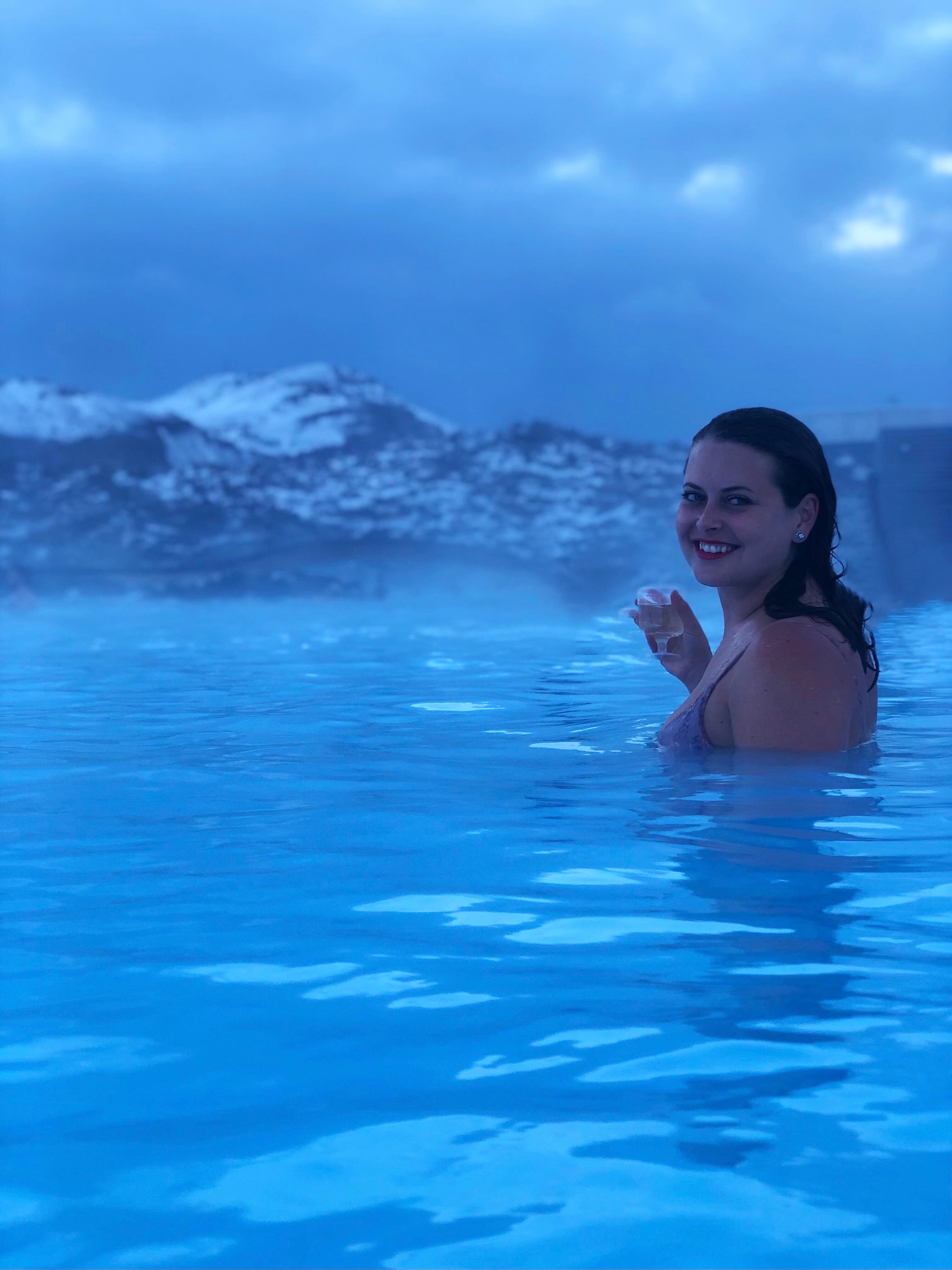 Advisor poses in a crystal-clear body of water with steam rising from the surface as mountains cascade toward the horizon at dusk.