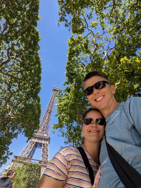 Monica and partner posing in front of the Eiffel Tower during the daytime.