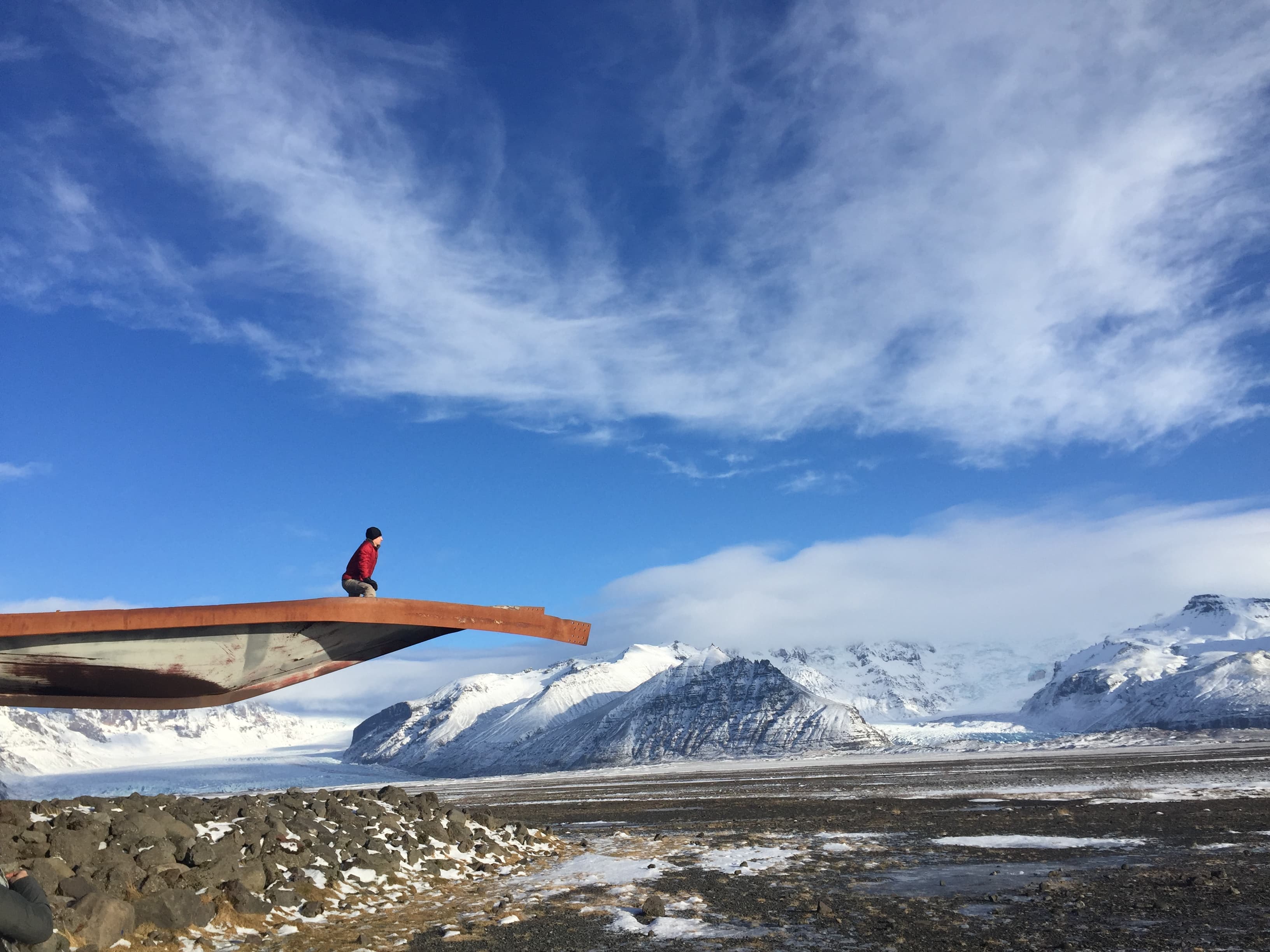 A person sitting on an overlook edge looking out onto a view of snowy mountains and rugged terrain.